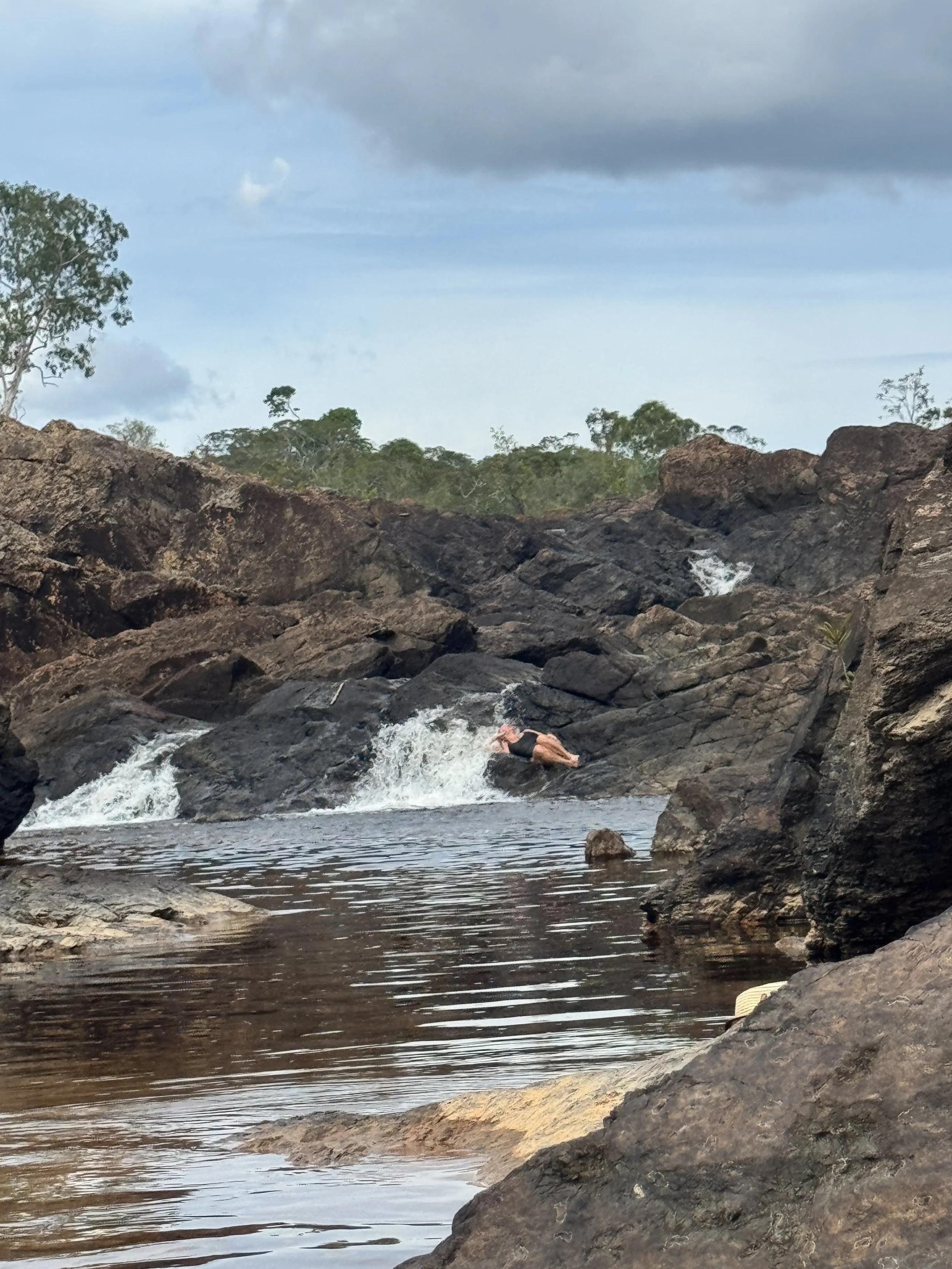 Person in a swimsuit lying on rocks near a small waterfall in a rocky river, surrounded by trees and cloudy sky.