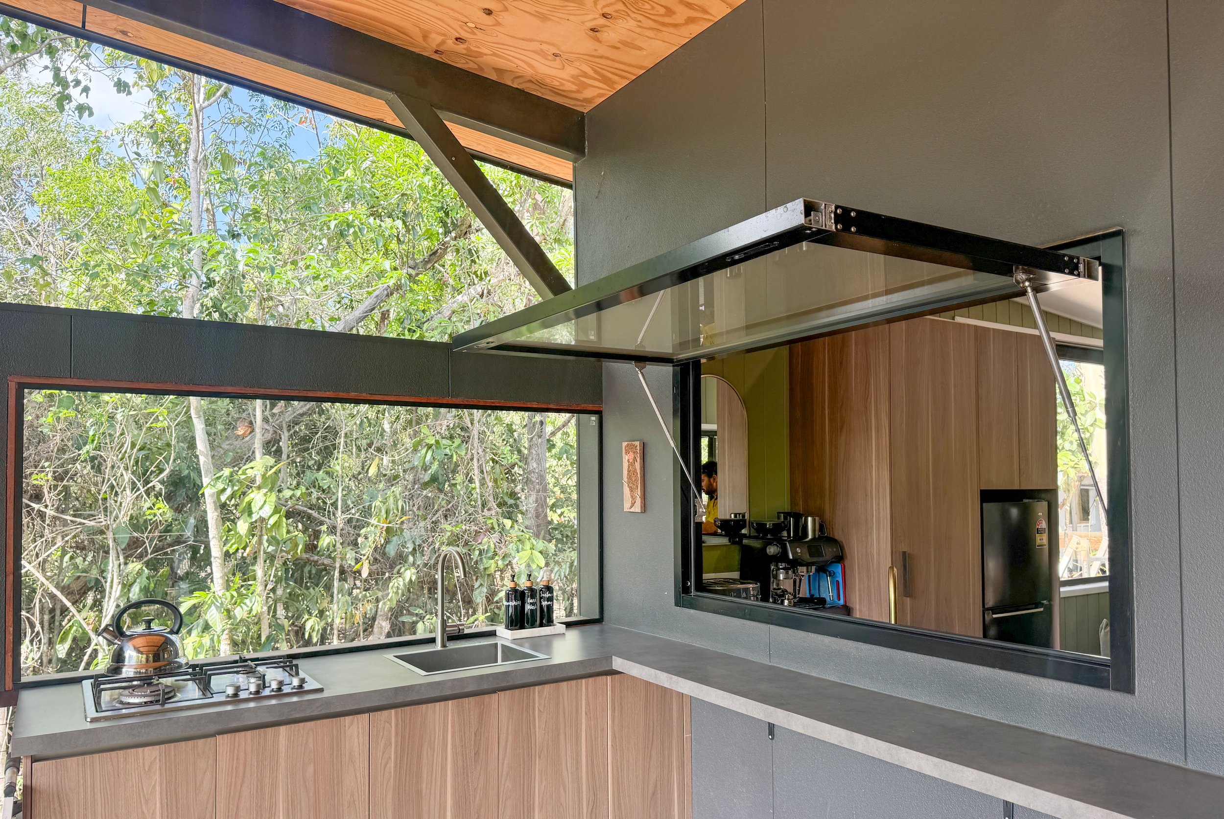 Modern kitchen with large windows showing trees outside, kitchen counter with sink, stovetop with kettle, and pass-through window to another room with kitchen appliances.