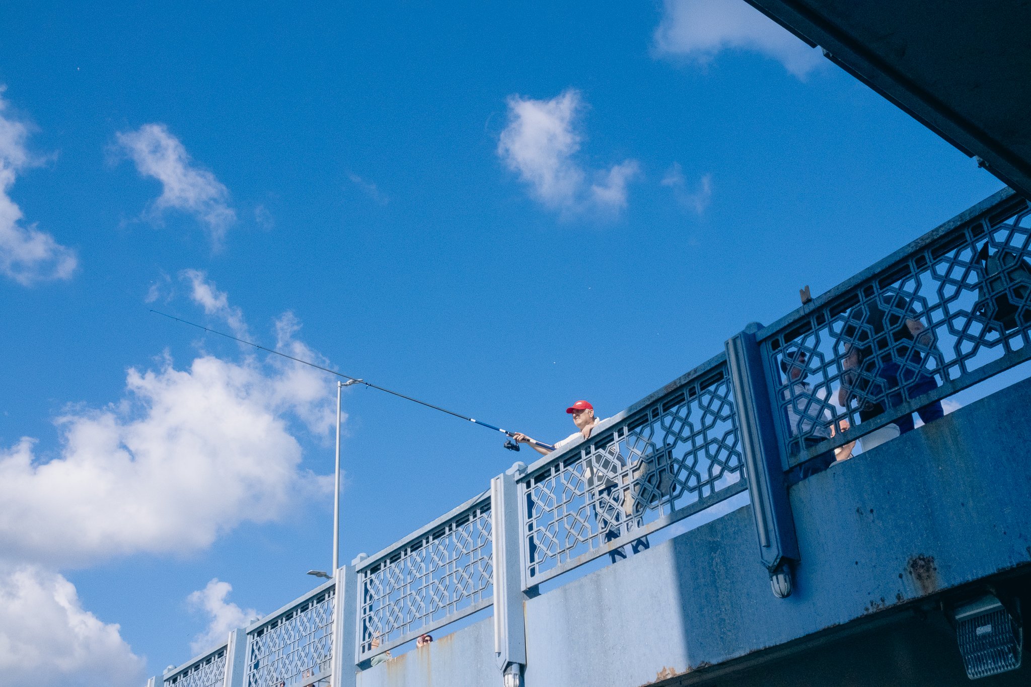 Person wearing a red cap fishing off a bridge with a clear blue sky and scattered clouds in the background.