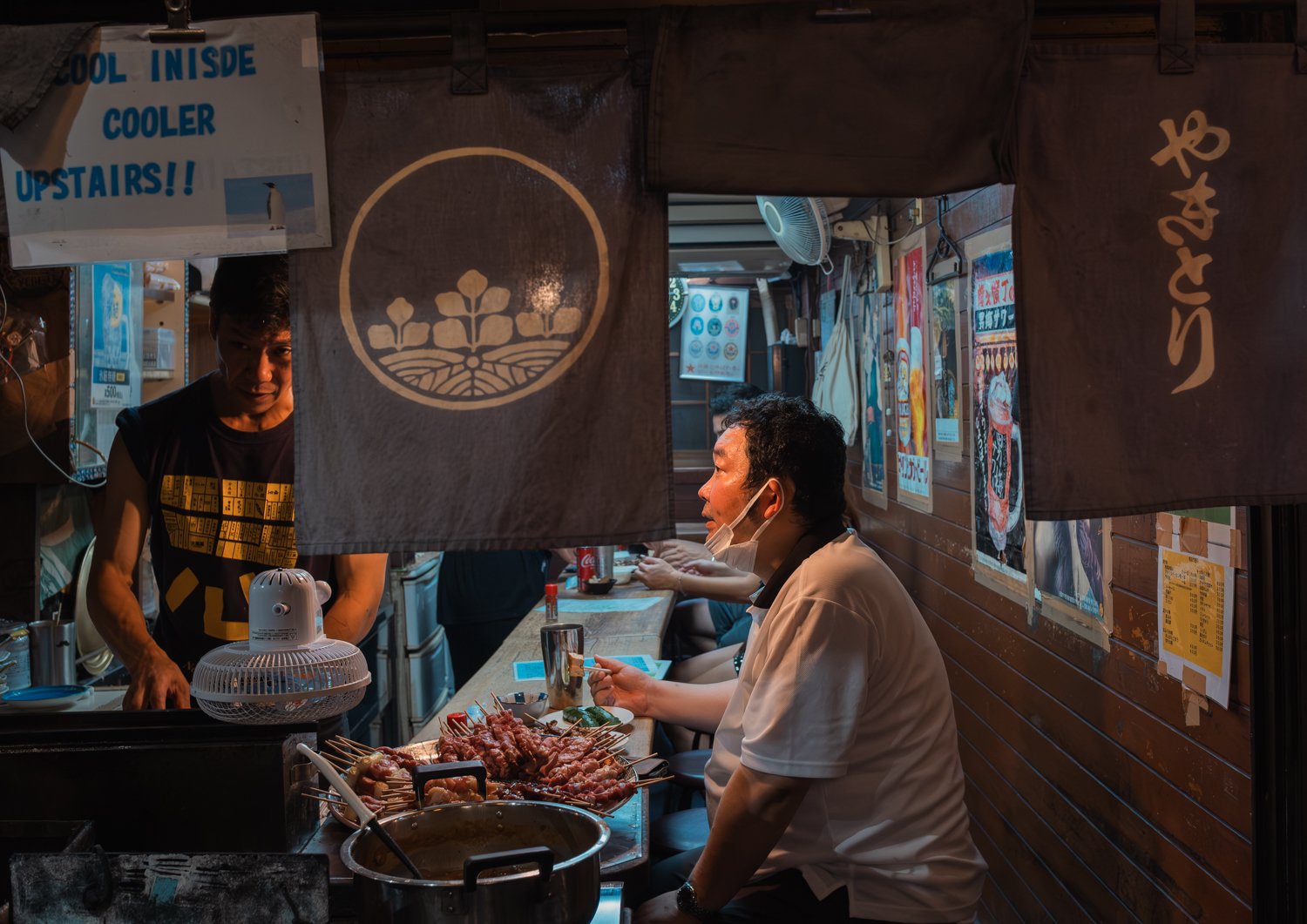 People sitting at a kitchen counter with skewers of grilled meat, with a chef preparing food behind the counter in a small restaurant or food stall.