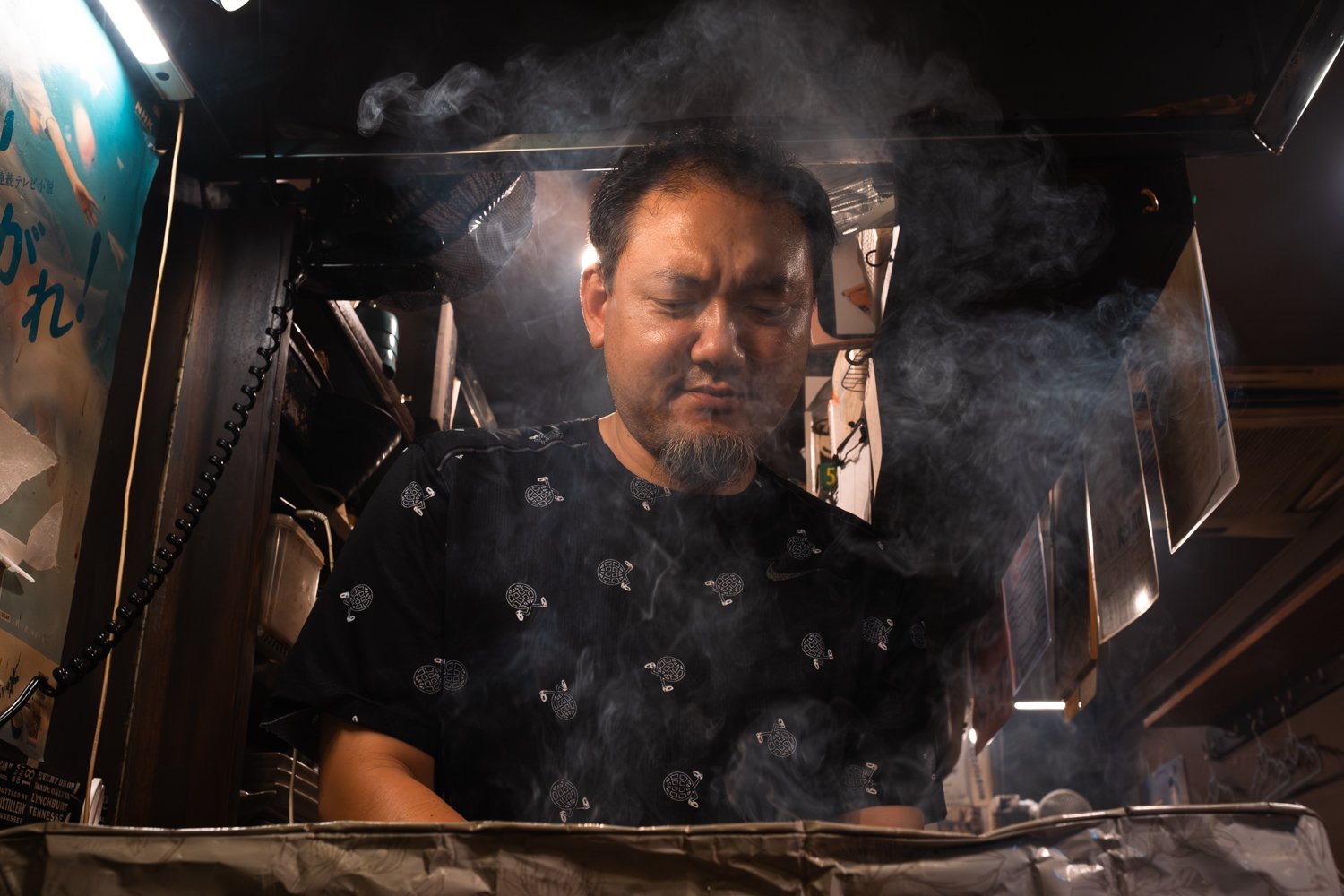 A man standing behind a counter, surrounded by smoke, with a focused expression, in a kitchen or food stall setting, with various kitchen tools and menus in the background.
