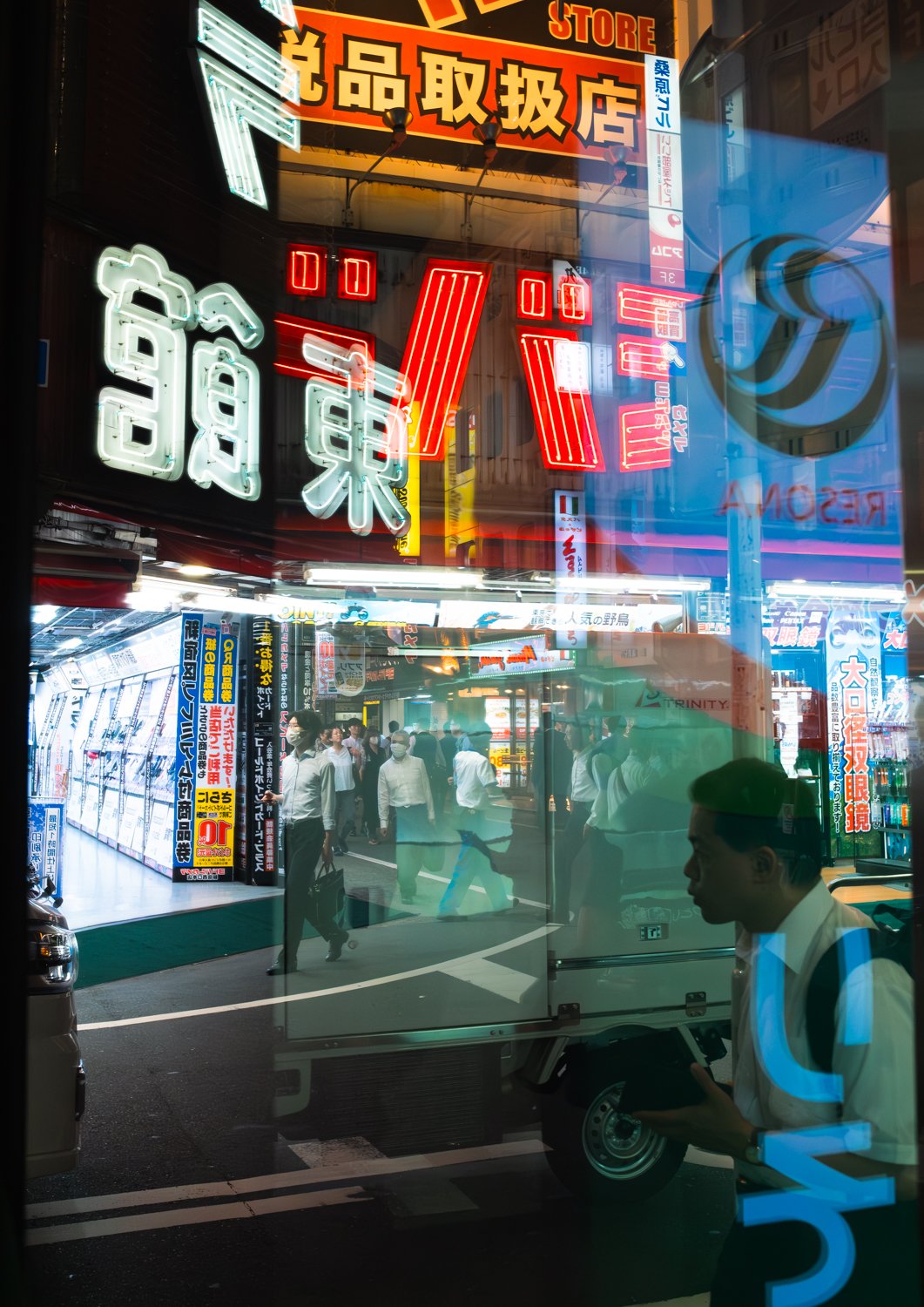 Night view of a busy street scene in Japan with neon signs, people walking, and a man wearing a mask holding a tablet reflections of lights and signs in a window