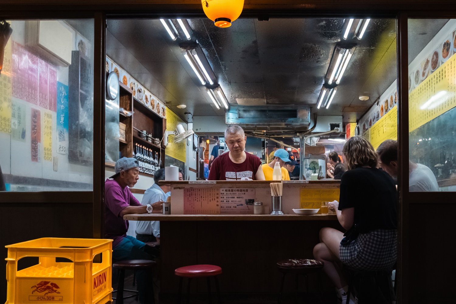 View inside a small Japanese ramen restaurant with customers seated at the counter and a chef preparing food behind the counter. Yellow and pink menus are visible on the walls, and a yellow crate with the Kirin beer logo is in the foreground.