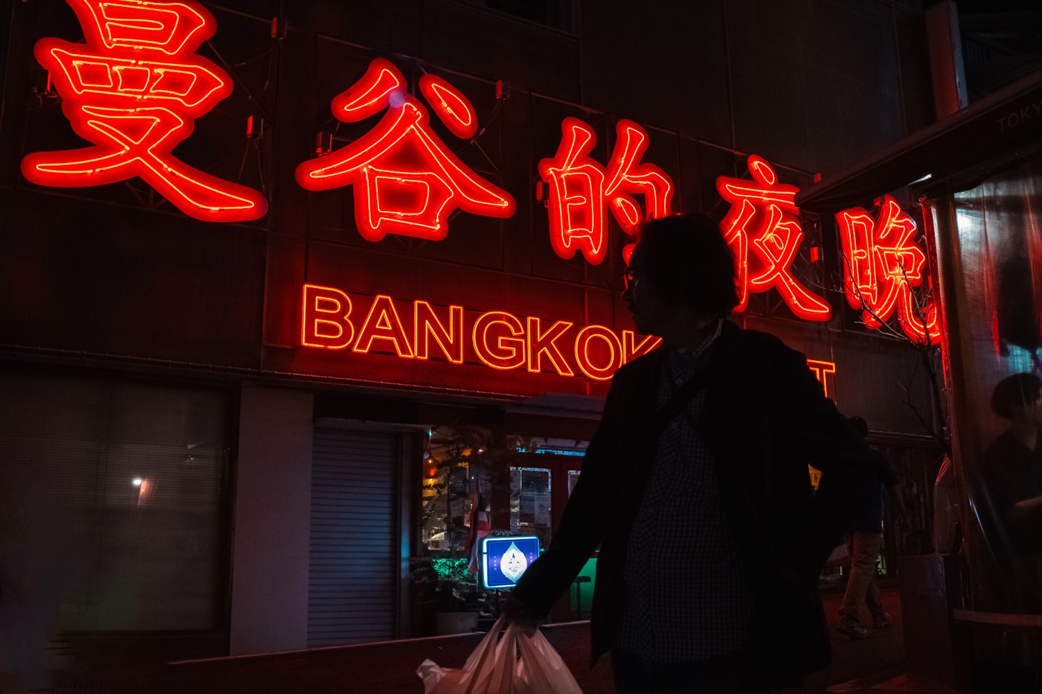 A silhouette of a person holding a plastic bag standing in front of a neon sign that reads 'BANGKOK'. The sign has Chinese characters above the English text and is illuminated in red. The scene is dark, with some reflection visible on a window behind