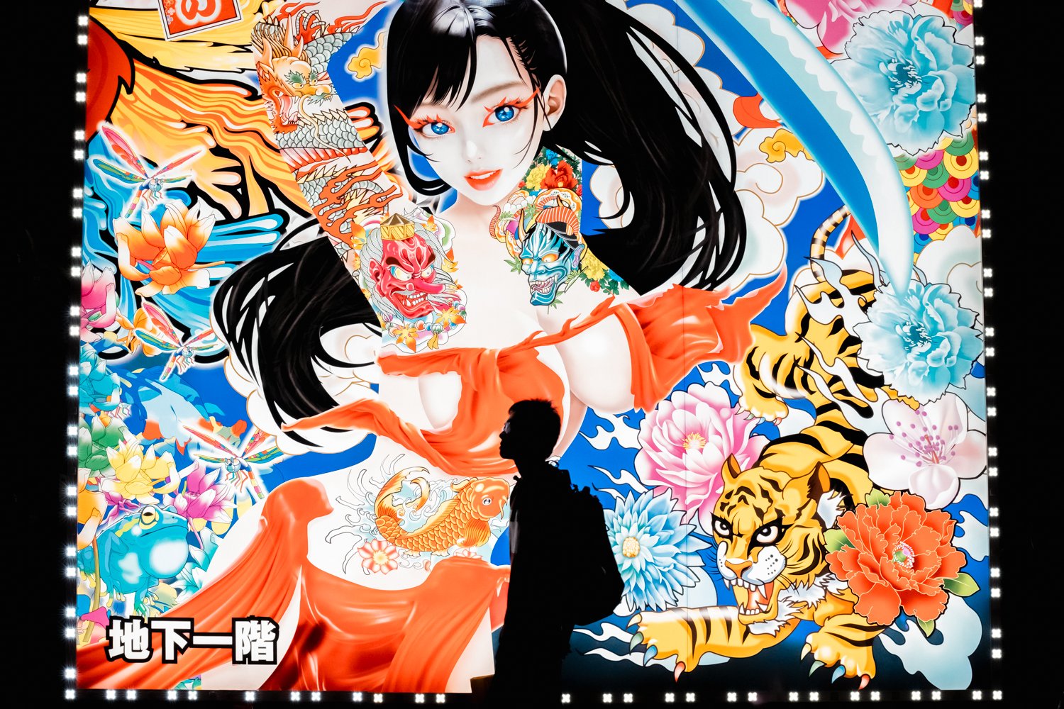 Silhouette of a person standing in front of a large, colorful tattoo-style mural featuring a woman with long black hair, blue eyes, and tattoos of traditional Japanese masks, flowers, a tiger, a dragon, and other mystical symbols.