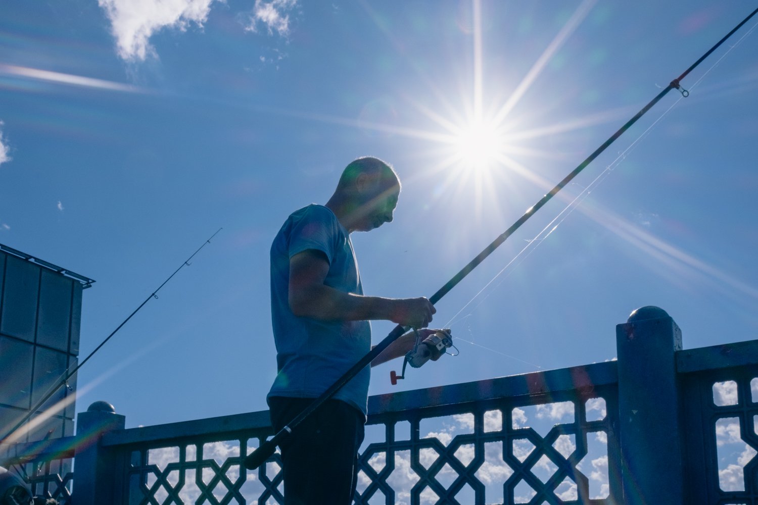 Man standing by a fence, fishing with a rod, under a bright sun in a clear blue sky.
