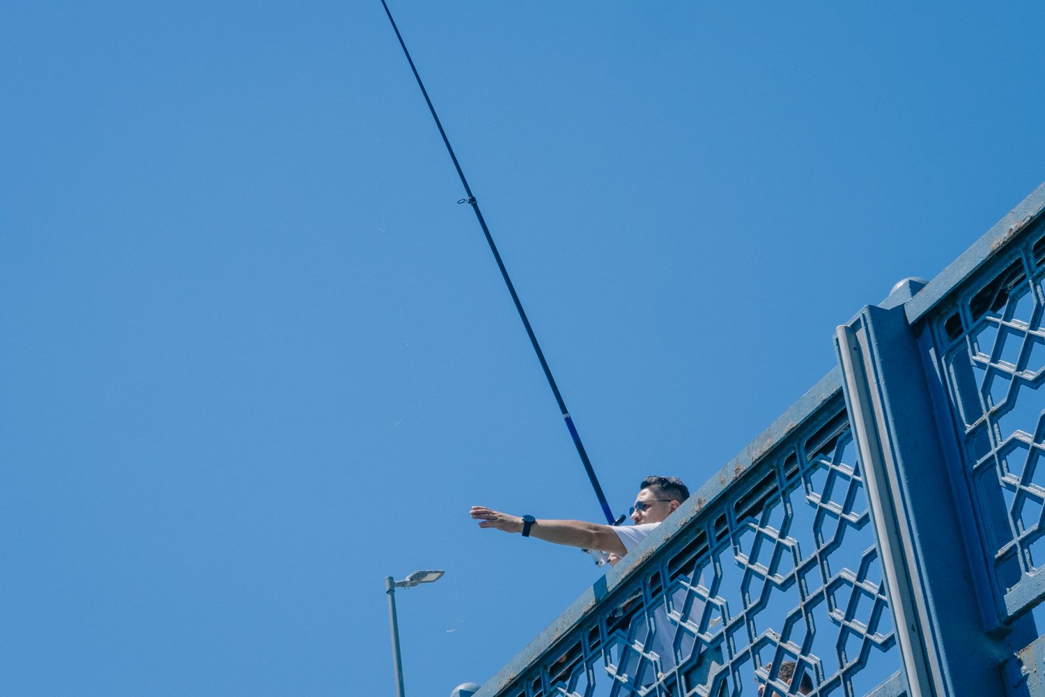 Man standing on a blue decorative bridge reaching out with his right arm, against a clear blue sky, with street lights and a fishing rod.