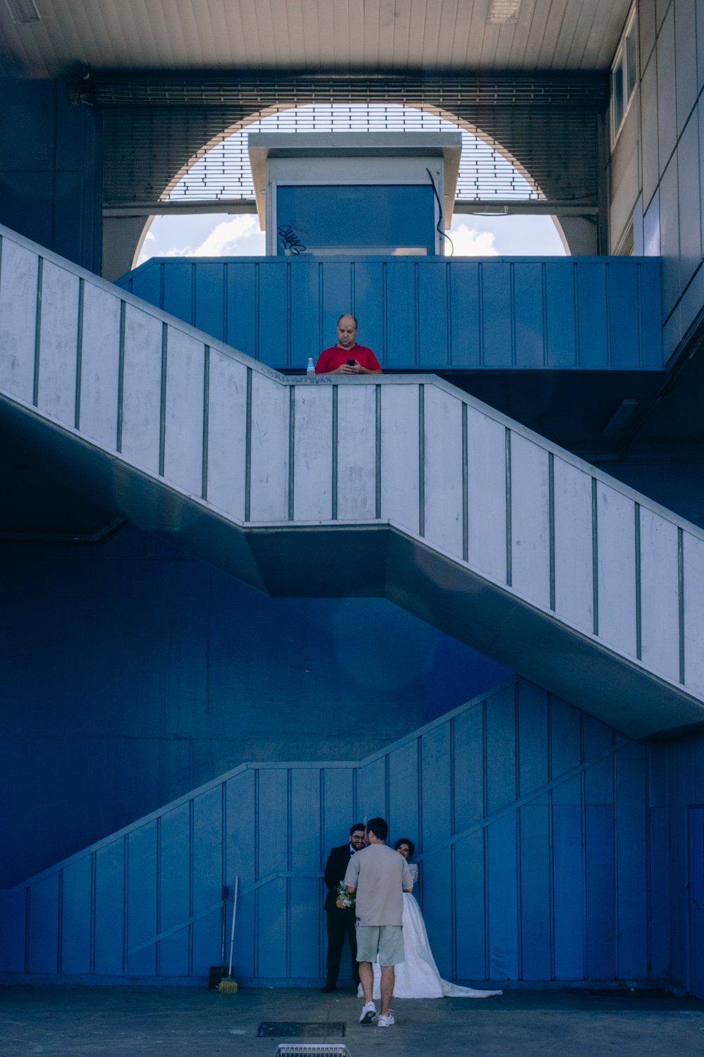 A man in a white shirt and shorts is taking a photo of a couple in formal attire, with the woman wearing a white dress, in front of a blue architectural staircase. There is another person above, dressed in a red shirt, looking at their phone.