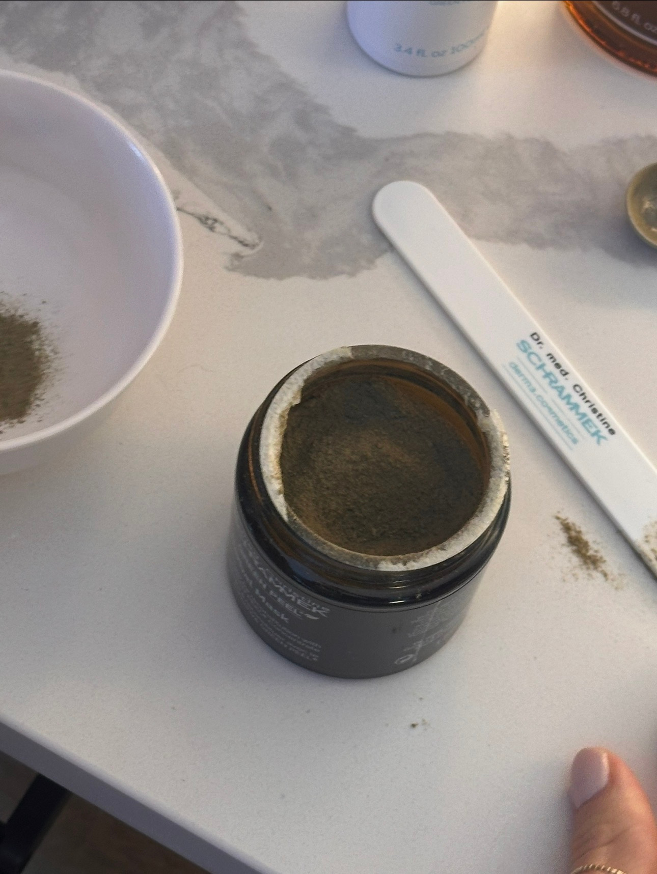Open jar of green powder supplement on a white table with a spoon labeled 'by me, online Schrammek' nearby, along with a bowl of green powder and other supplement containers.