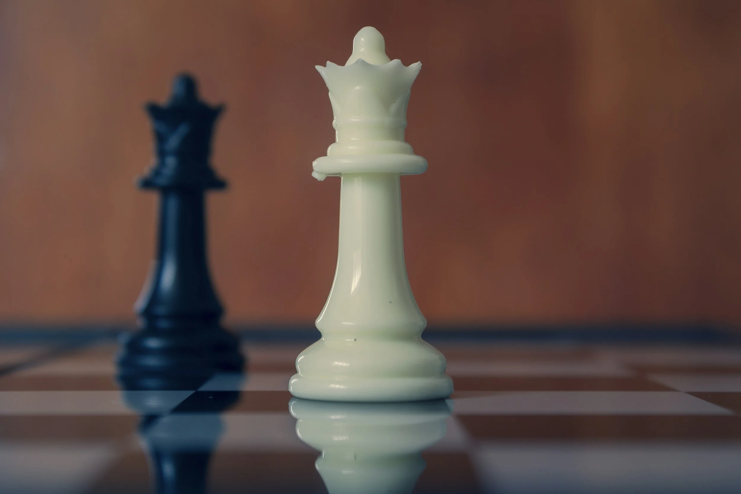 Close-up of a white queen chess piece on a wooden chessboard with a blurred black queen in the background.