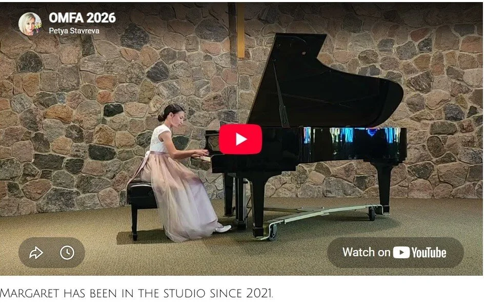 A young girl with dark hair, wearing a white top and a long pink skirt, playing a grand piano in a room with a stone wall background.