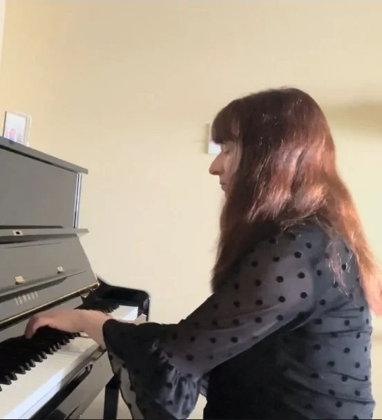 A woman with long brown hair playing a piano in a room with beige walls.