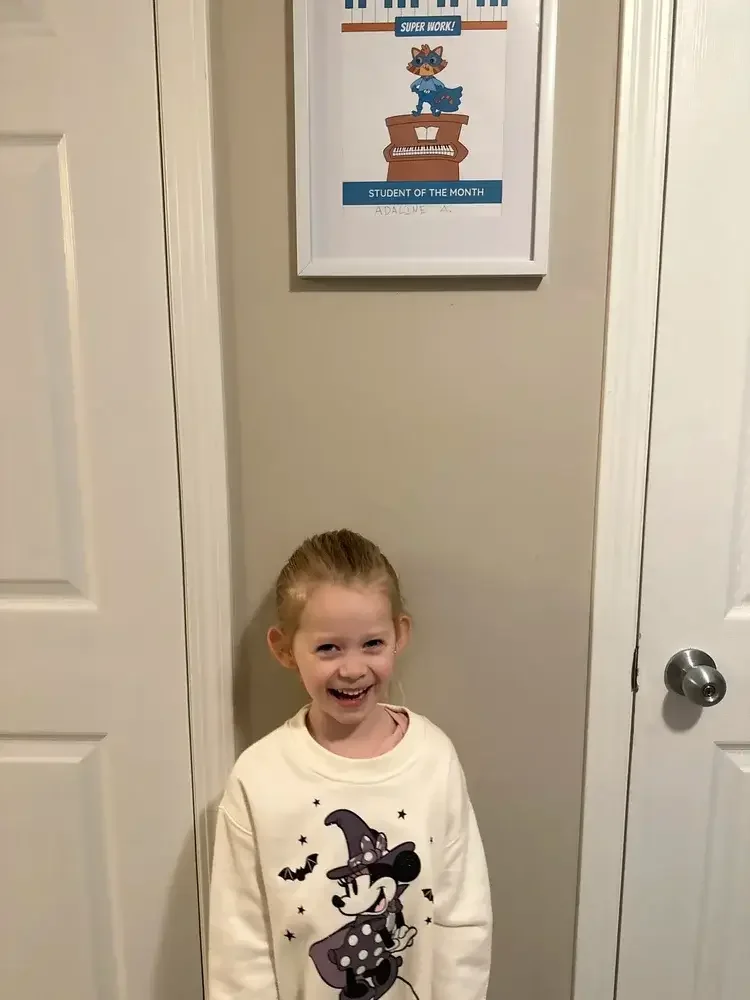 A young girl with red hair pulled back into a ponytail, smiling and standing in front of a beige wall with a framed certificate above her head. The certificate is titled "Student of the Month" featuring a cartoon raccoon playing a piano, with a label that says "Super Work!"
