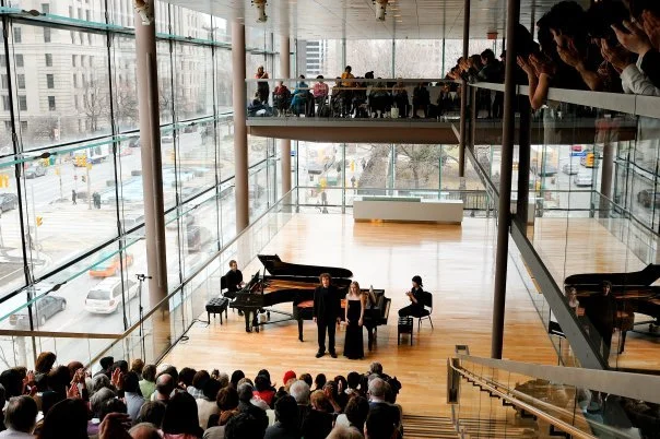 Musicians performing on stage in a modern, glass-walled concert hall, with an audience seated on the main floor and mezzanine level.