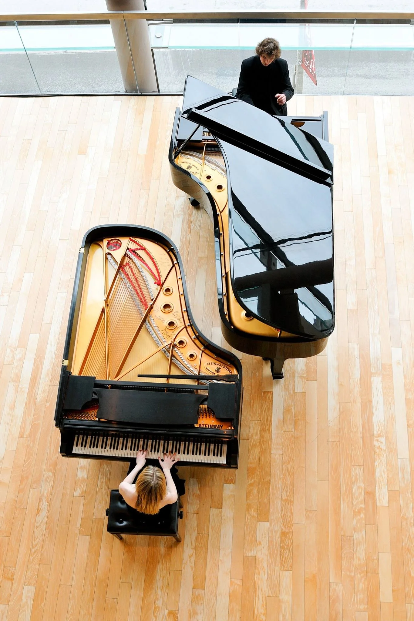 Top view of a performance space with a woman playing a grand piano and a man standing behind it. The grand piano is open, showing its strings and mechanical parts, with another closed black piano nearby. The room has light wood flooring and a large window in the background.