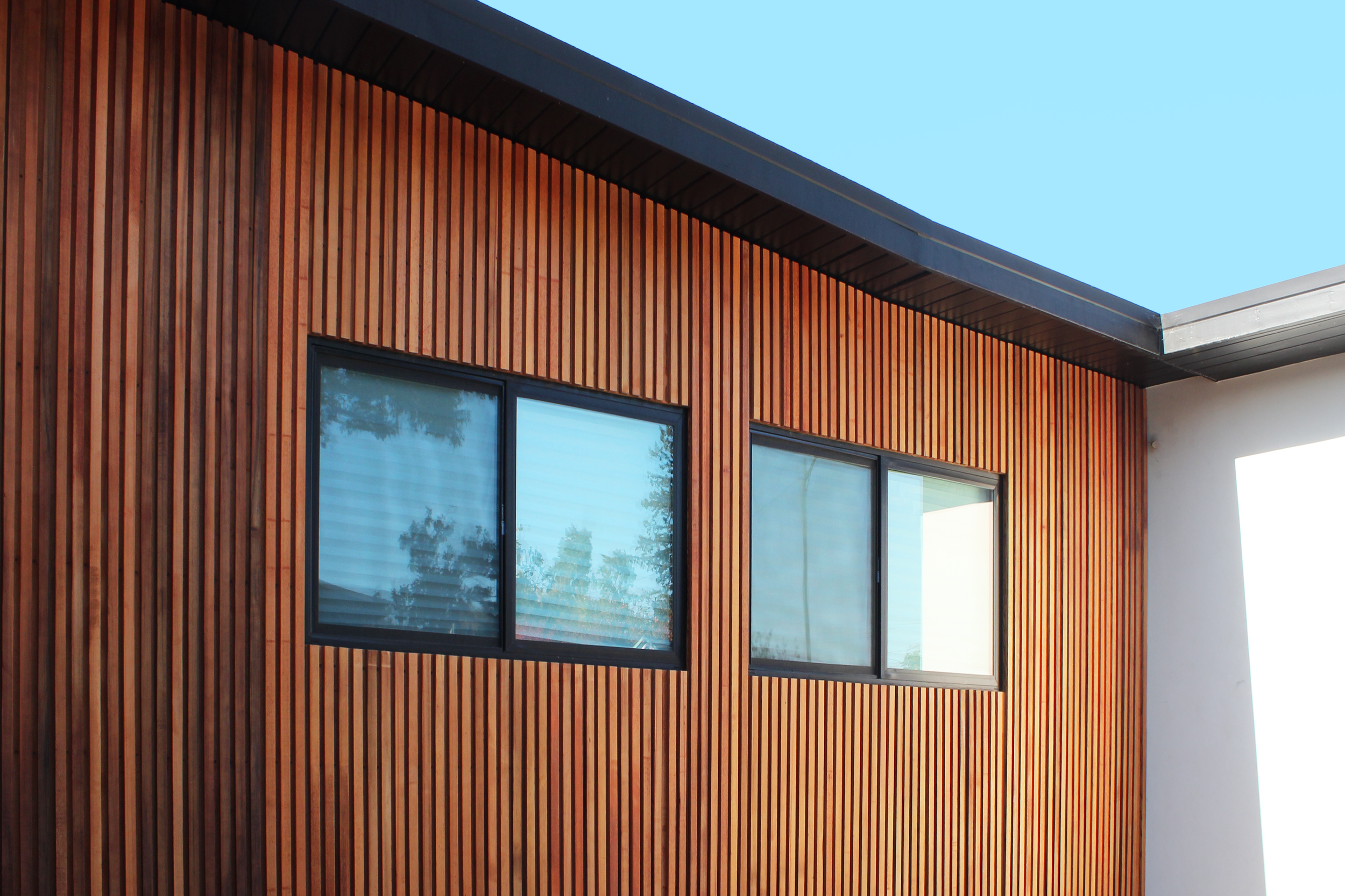 Exterior view of a modern building with a wooden slat facade, two large windows, and a sloped roof against a clear blue sky.