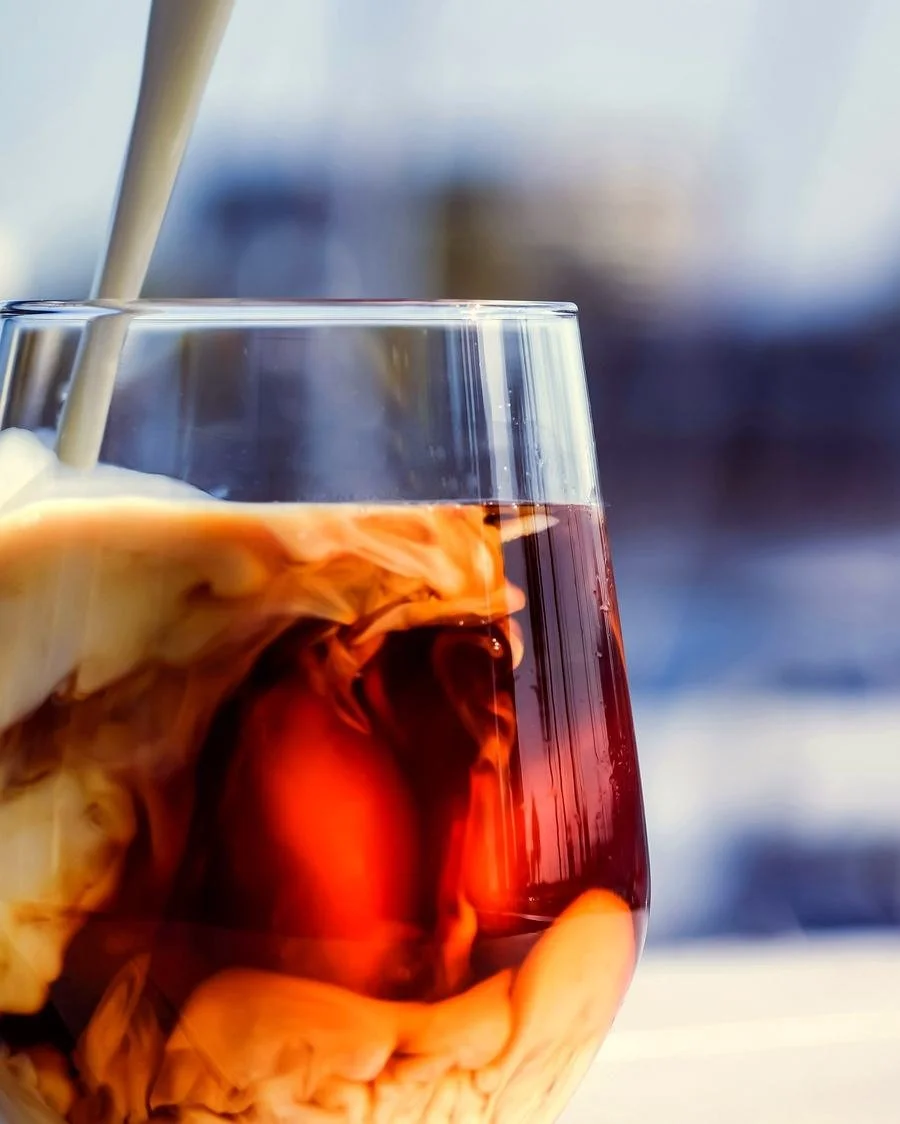 A glass of iced tea with ice cubes and cream being poured in, creating swirling patterns.
