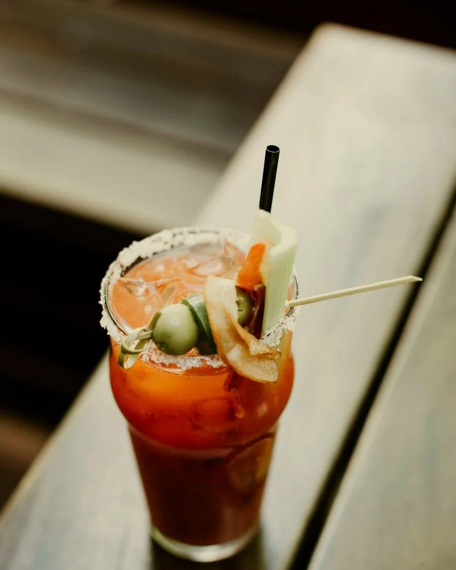 A tall glass of Bloody Mary cocktail garnished with celery, lime, and a slice of bread on a skewer, with a salted rim and black straw
