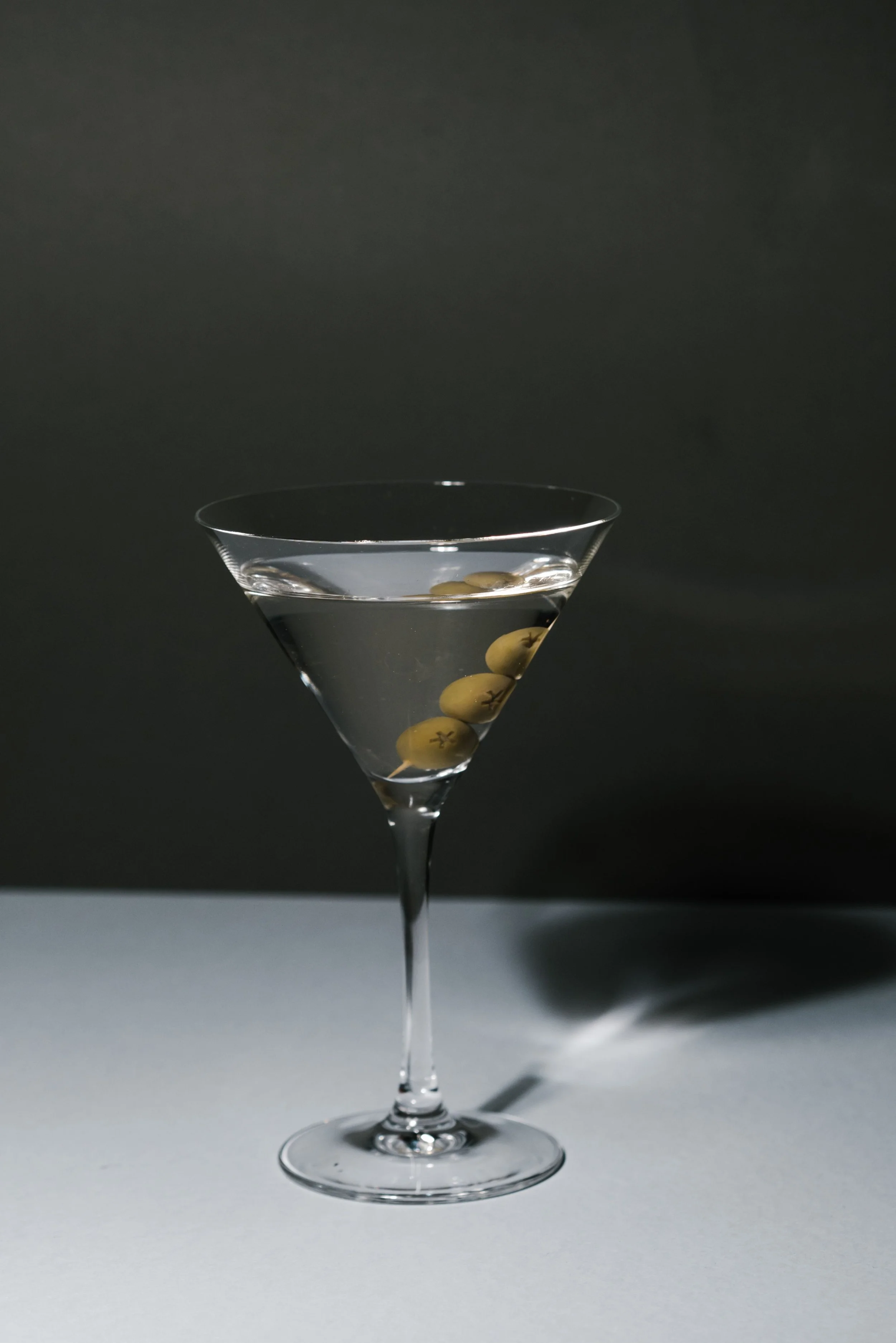 A martini glass filled with clear liquid and three green olives on a skewer, set against a dark background.