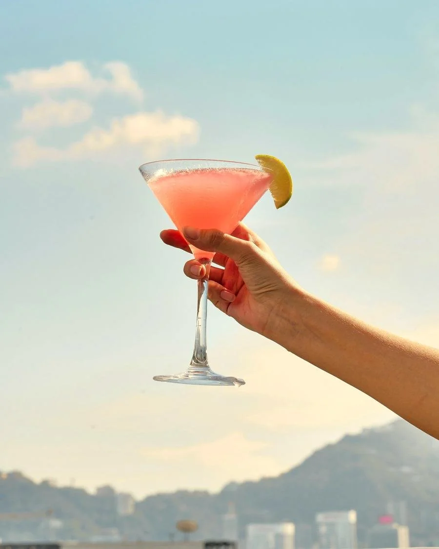 A person holding a pink cocktail in a martini glass with a lemon wedge garnish, set against a daytime sky with some clouds and distant mountains.