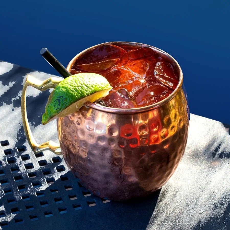 Copper Moscow Mule mug filled with iced beverage, garnished with a lime wedge and black straw, resting on a outdoor table with blue sky background.
