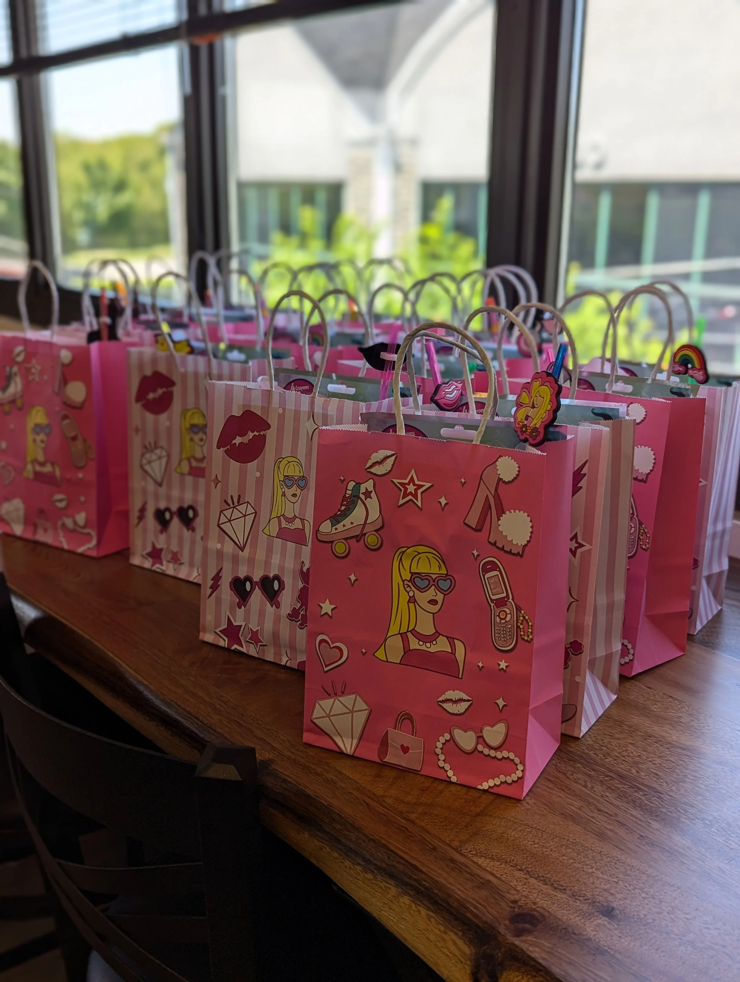 Pink gift bags decorated with illustrations of a girl with sunglasses, hearts, lips, a diamond, a roller skate, a handbag, a cellphone, and other fashion-themed icons, arranged on a wooden surface near a window.