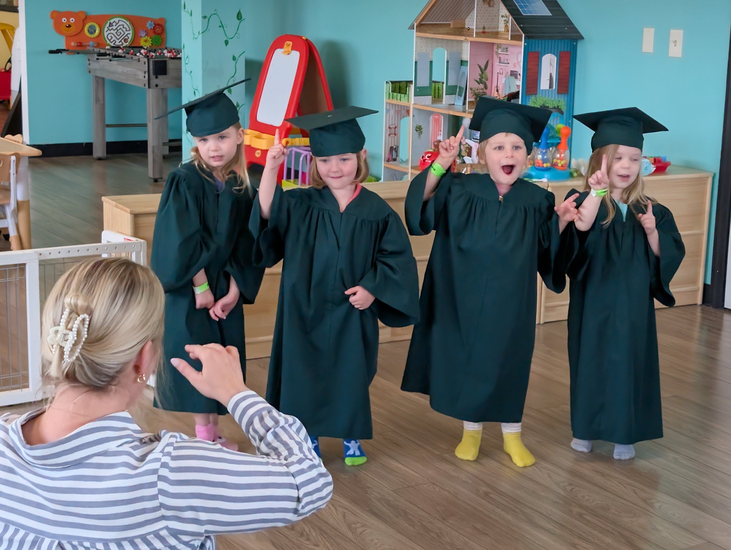 Four children wearing black graduation caps and gowns standing inside a colorful playroom, with a woman sitting in front of them and raising her hand.