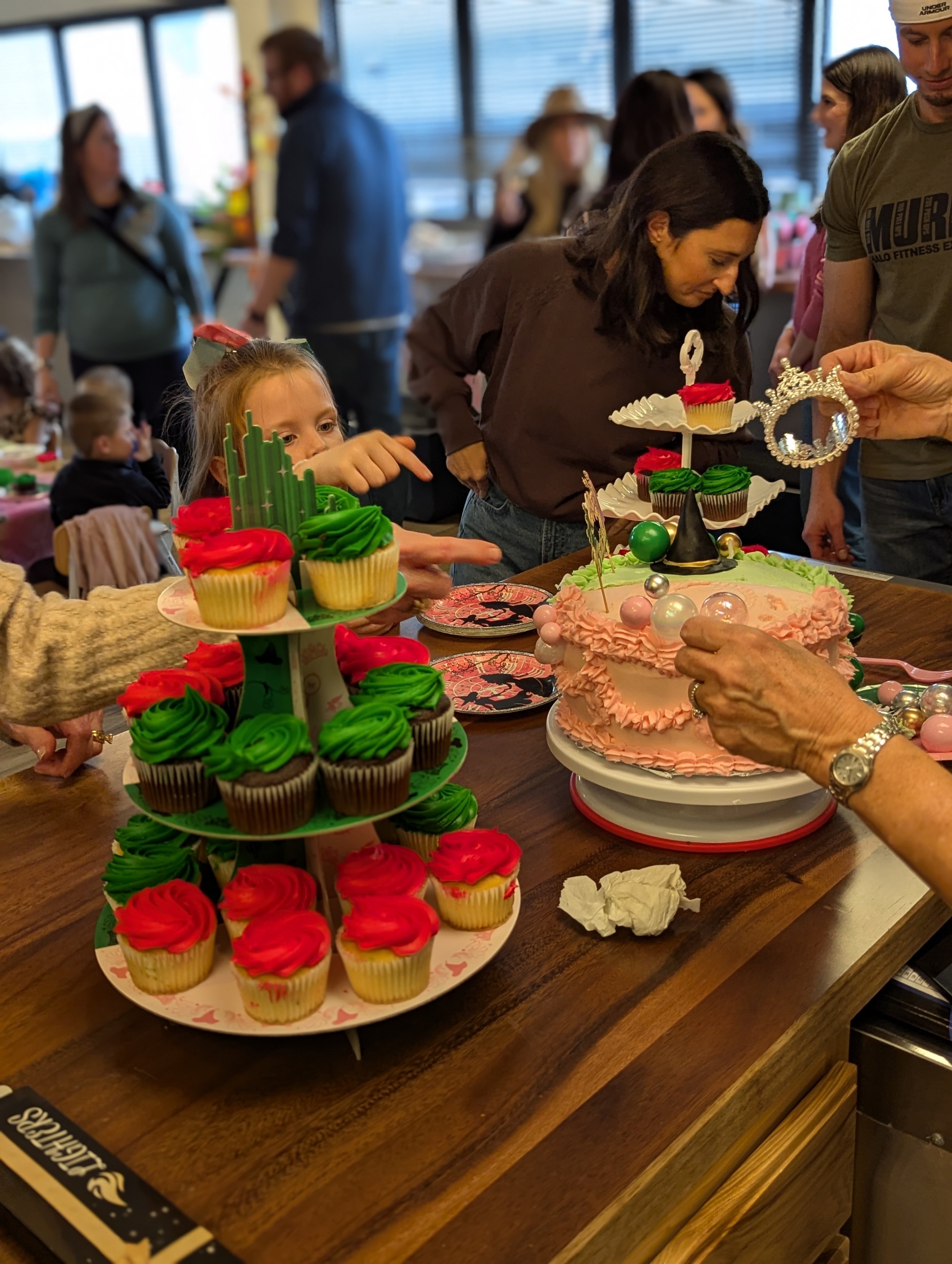 People gathered around a table celebrating a birthday with cupcakes and a decorated cake, in a cozy indoor setting.