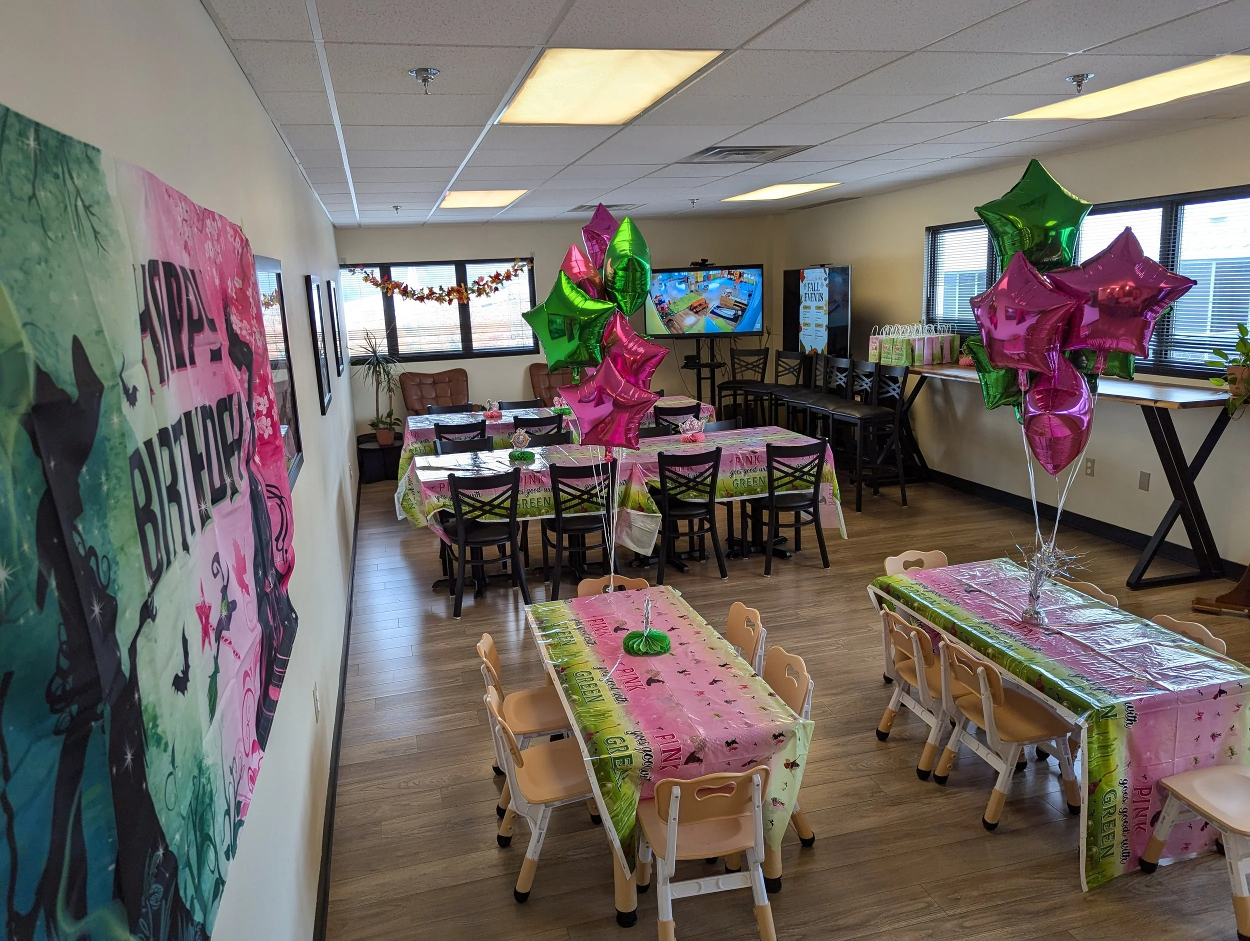 Party room decorated for a birthday celebration with pink and green balloons, birthday-themed tablecloths, and a spray of star-shaped balloons, with tables and chairs arranged for guests.