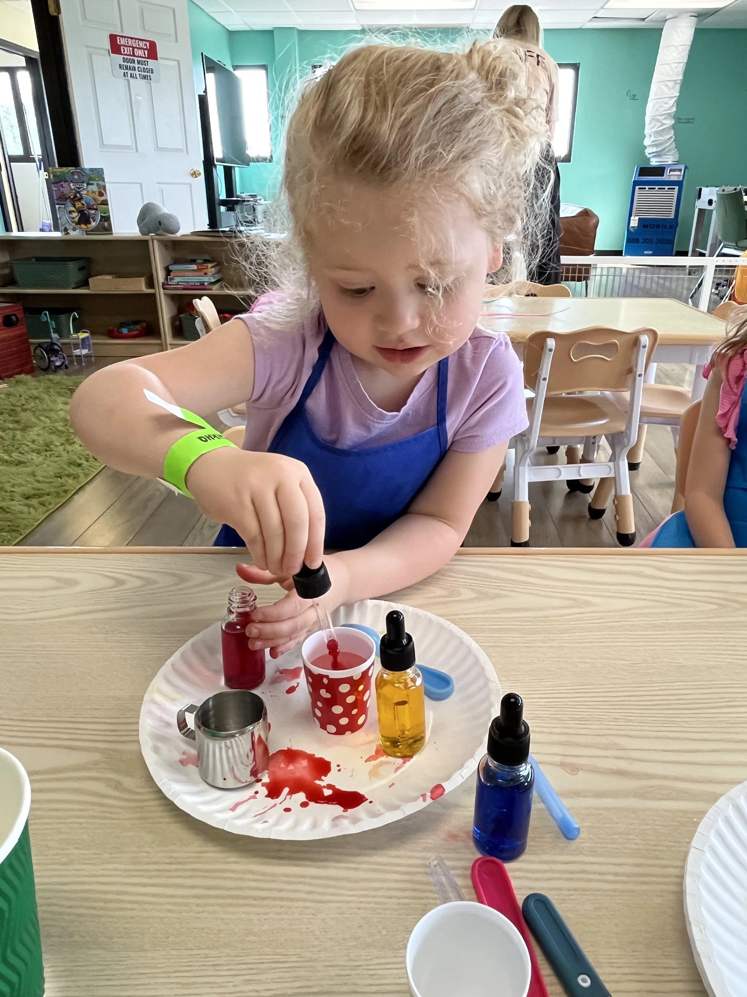 A young girl with curly blonde hair wearing a purple shirt and blue apron, engaged in a science activity with colored liquids in a classroom setting. She is holding a dropper over a red and white polka dot cup, with various bottles and utensils on a 