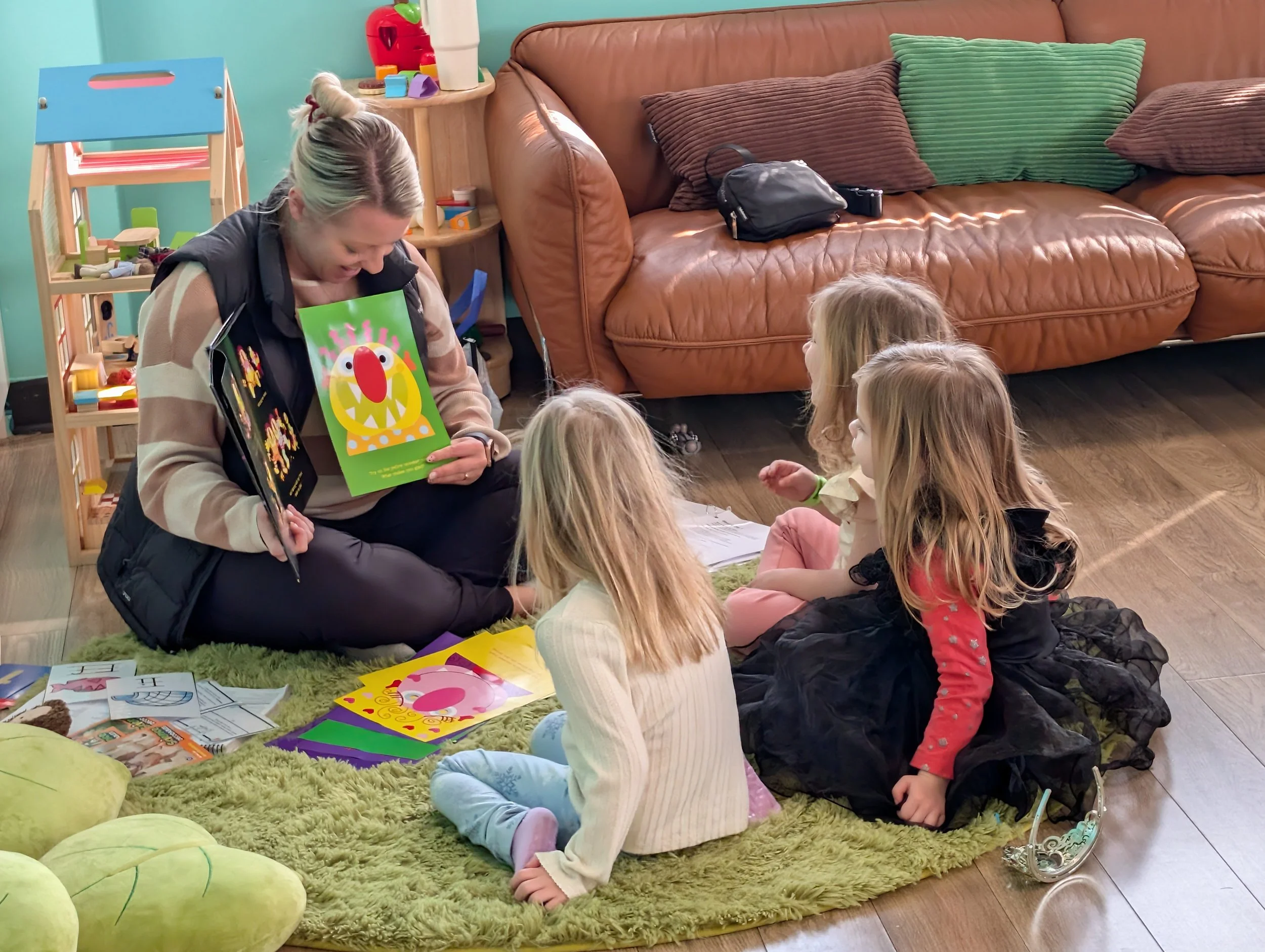 A woman and children sitting on a green rug, looking at colorful drawings and pictures. There's a brown leather couch with pillows in the background, a small round table, and a wooden shelf with toys.