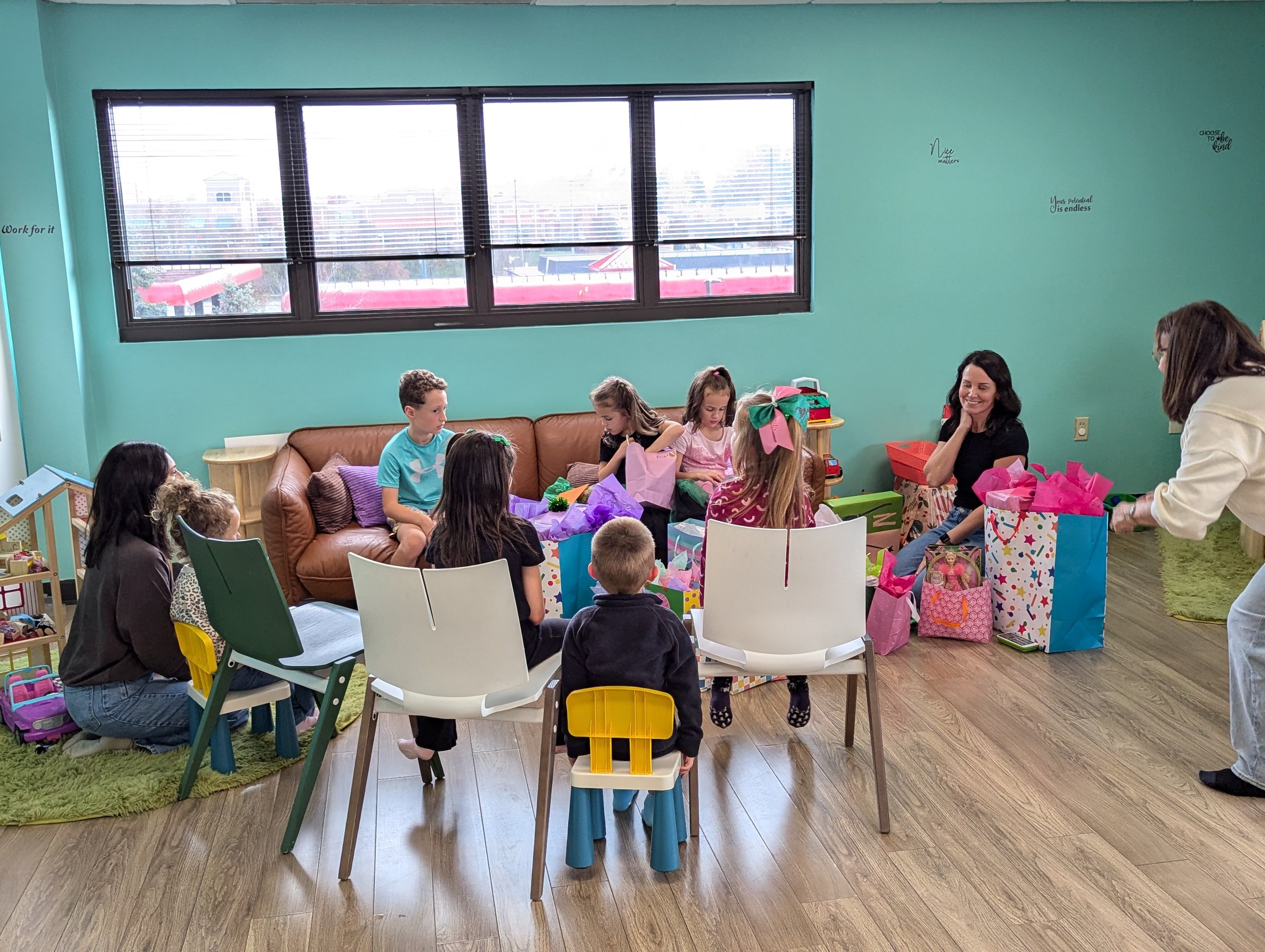 A birthday party with children opening presents in a room with mint green walls. There is a tan couch, a small table, and a mix of chairs. The children sit on or around the couch, surrounded by colorful gift bags and wrapping paper. Two women are org