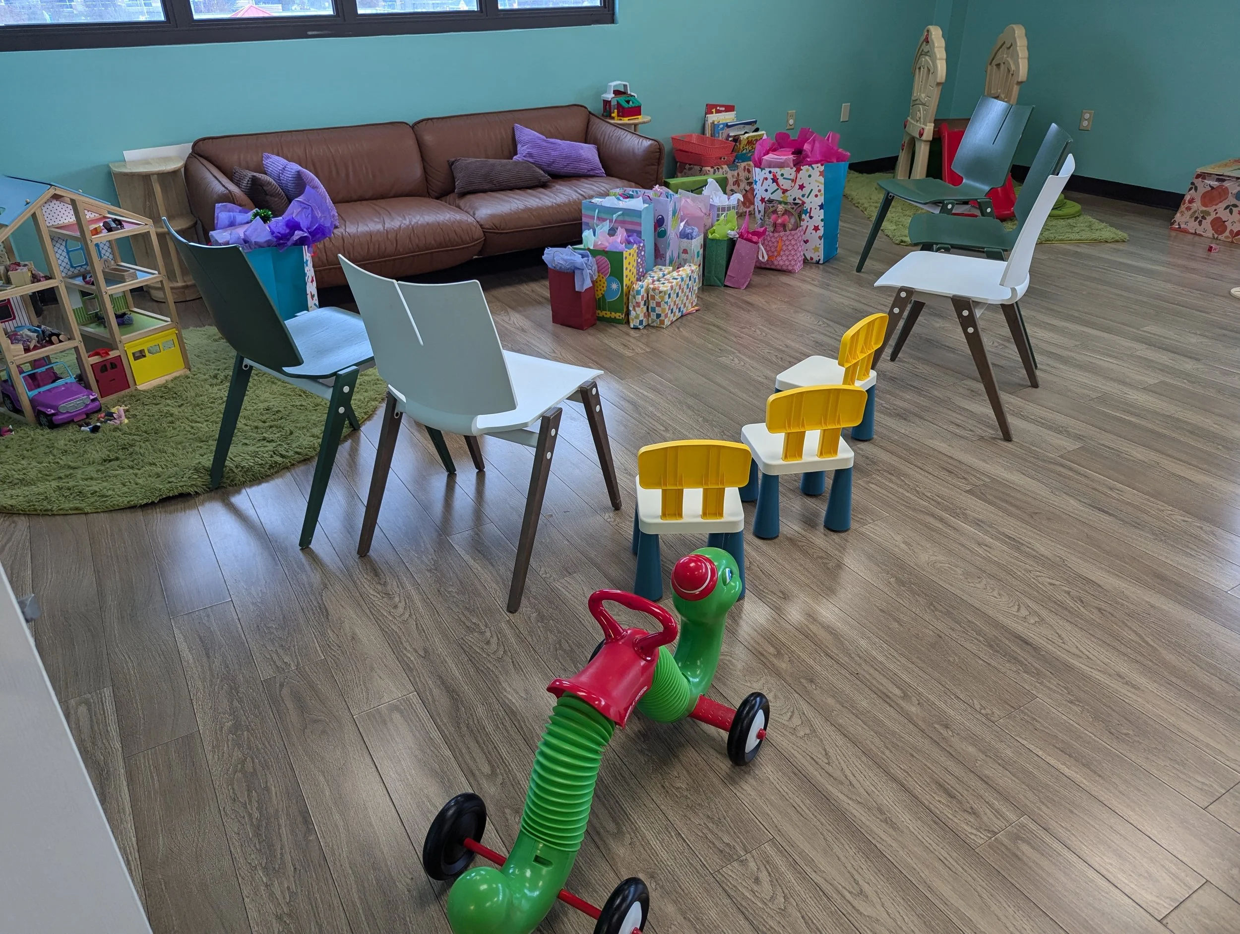 Room with a brown leather sofa, colorful children's toys and gifts, yellow and white children's chairs, and a green and red scooter on wooden floor.