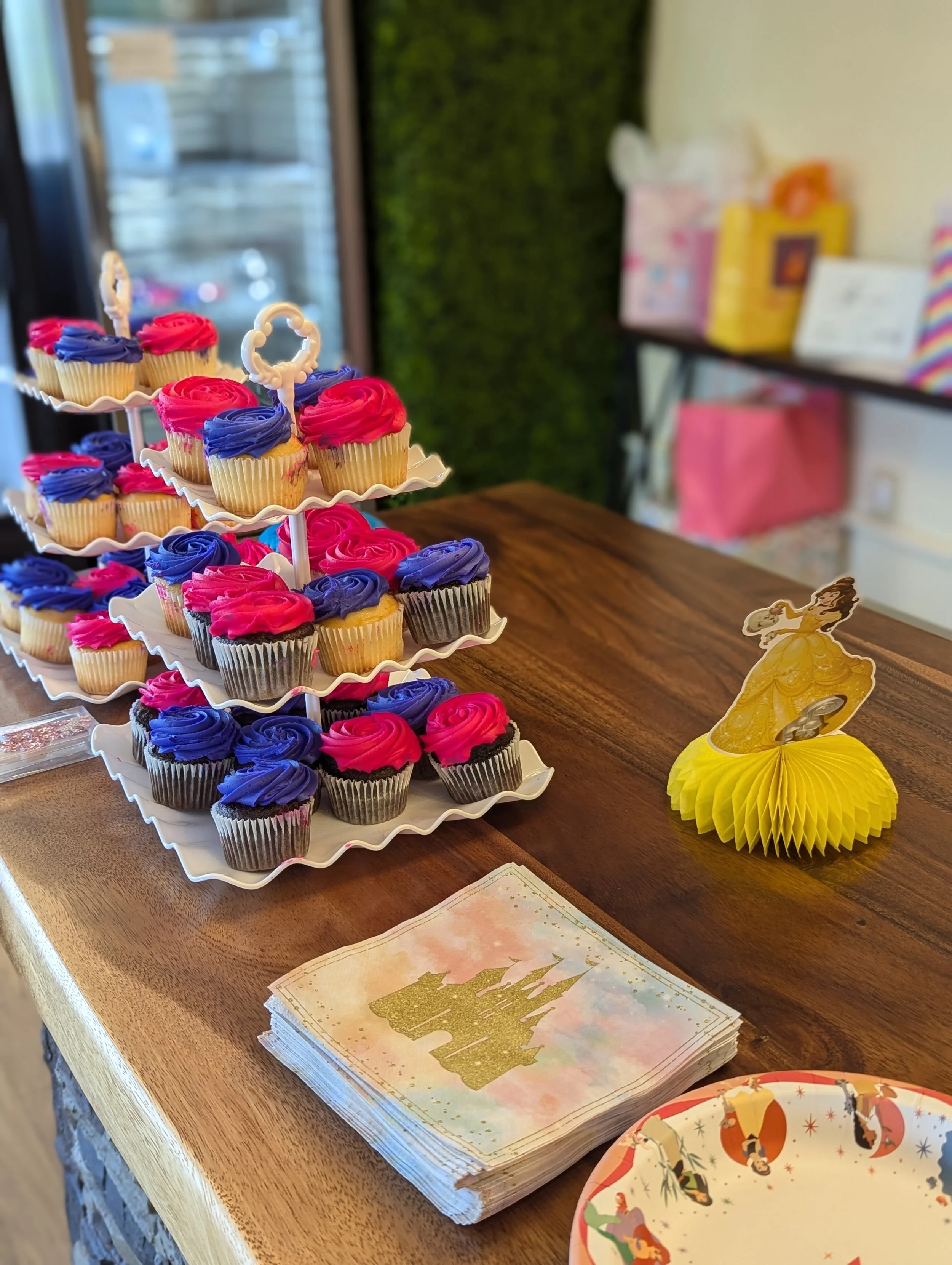 Colorful cupcakes with pink and blue frosting on tiered white stand, princess-themed party decorations, and a Disney princess figure on yellow paper decoration, on a wooden table.