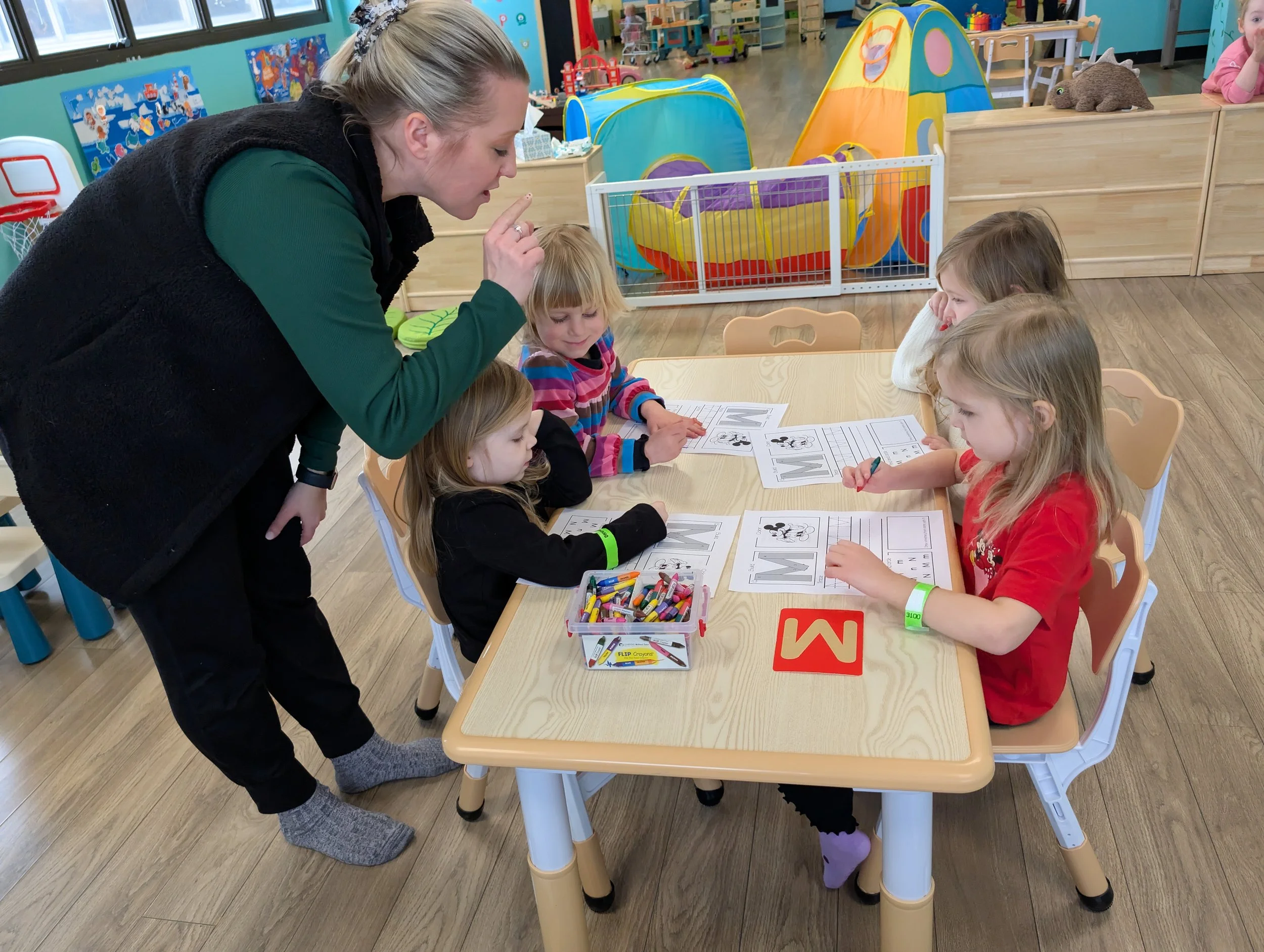 A teacher helping four young children with a worksheet at a table in a colorful classroom, with toys and decorations in the background.