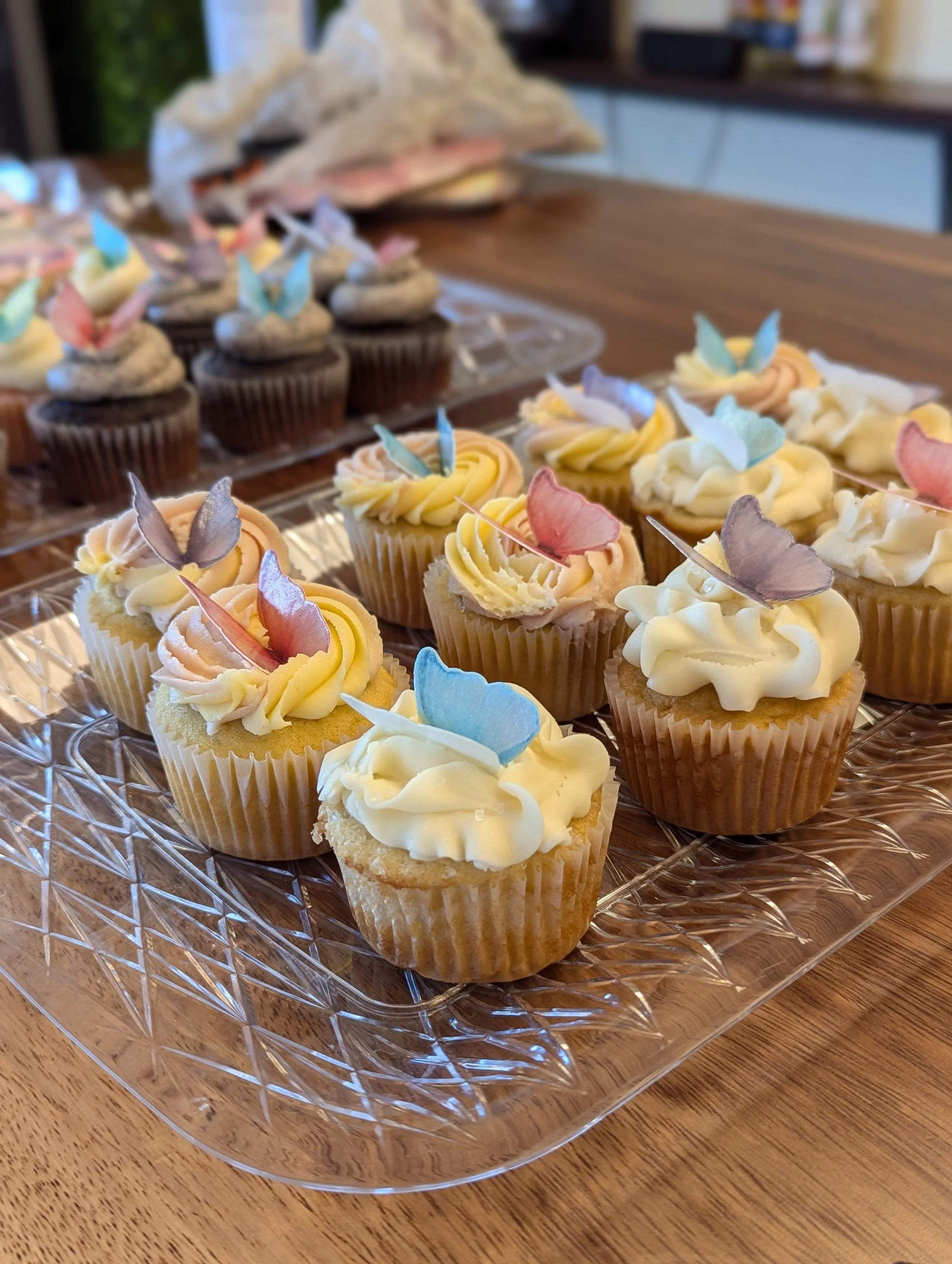 Assorted cupcakes decorated with pastel-colored butterfly and flower toppers on a glass tray.