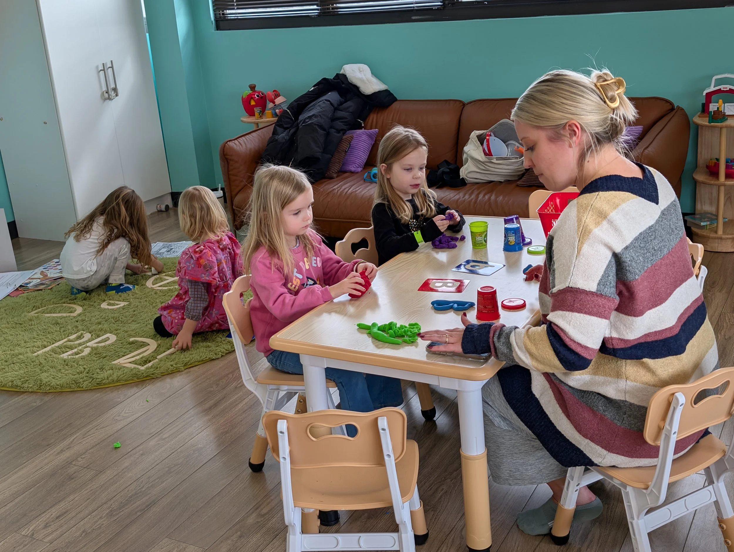A woman teaching a group of young girls in a classroom, with some girls sitting at a table playing with slime, while one girl is on the floor reading a book on a rug.
