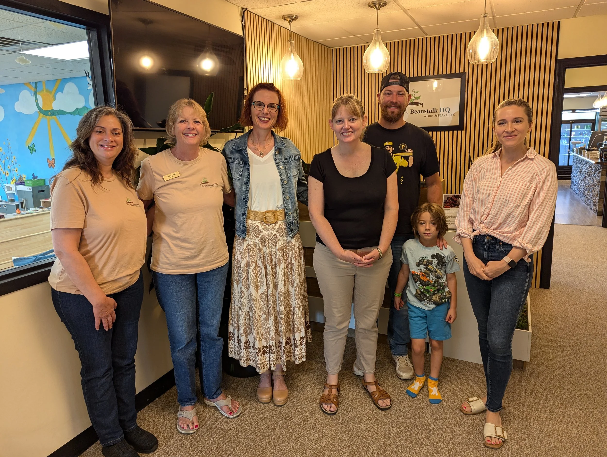 Group of seven people, including five women, one man, and a young girl, standing indoors at a workplace or café with wooden wall decor and a sign that says 'Beanstalk HQ Work & Play Café'.