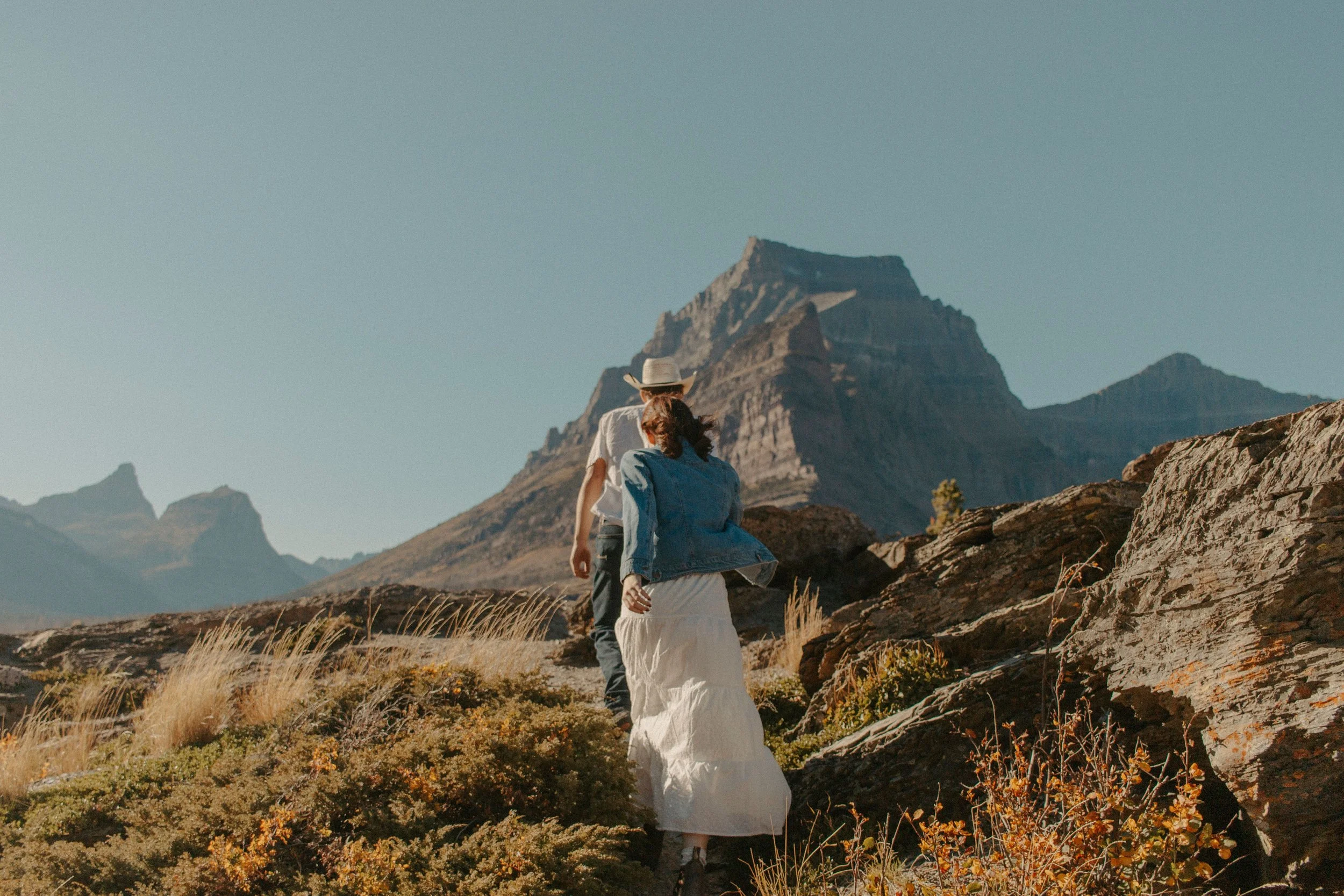 couple walks up cliff with mountains in the back