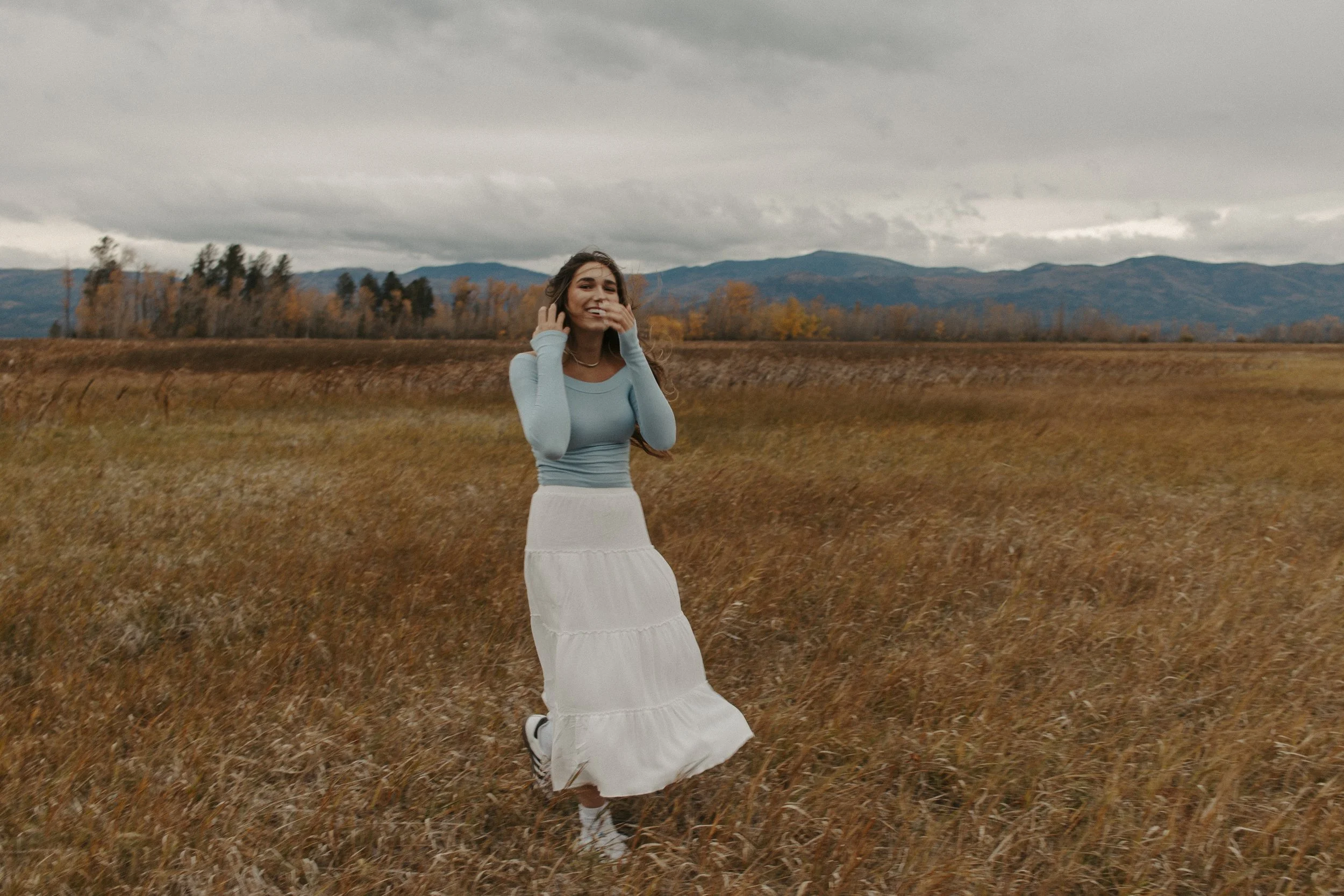 girl in long white skirt runs in field with mountains in the back