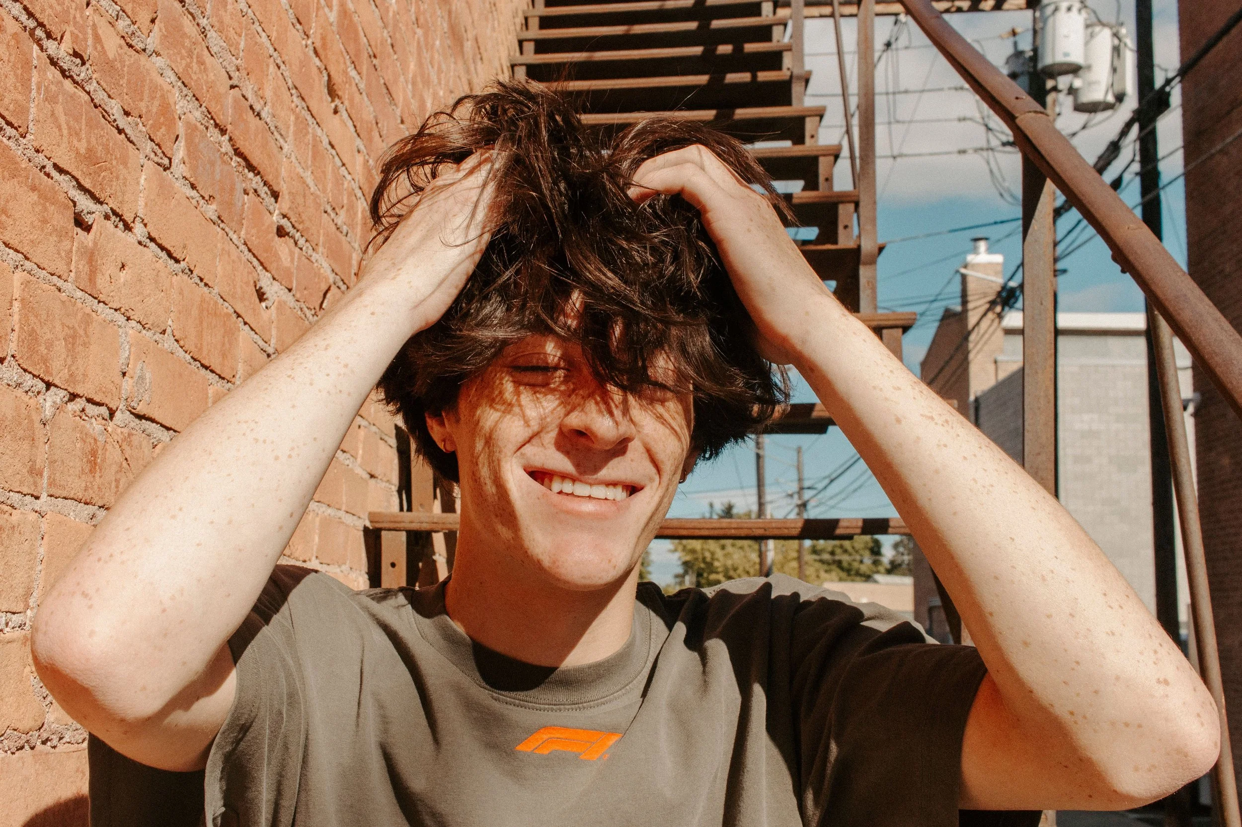 boy has his eyes closed while he fixes his hair on metal staircase outside