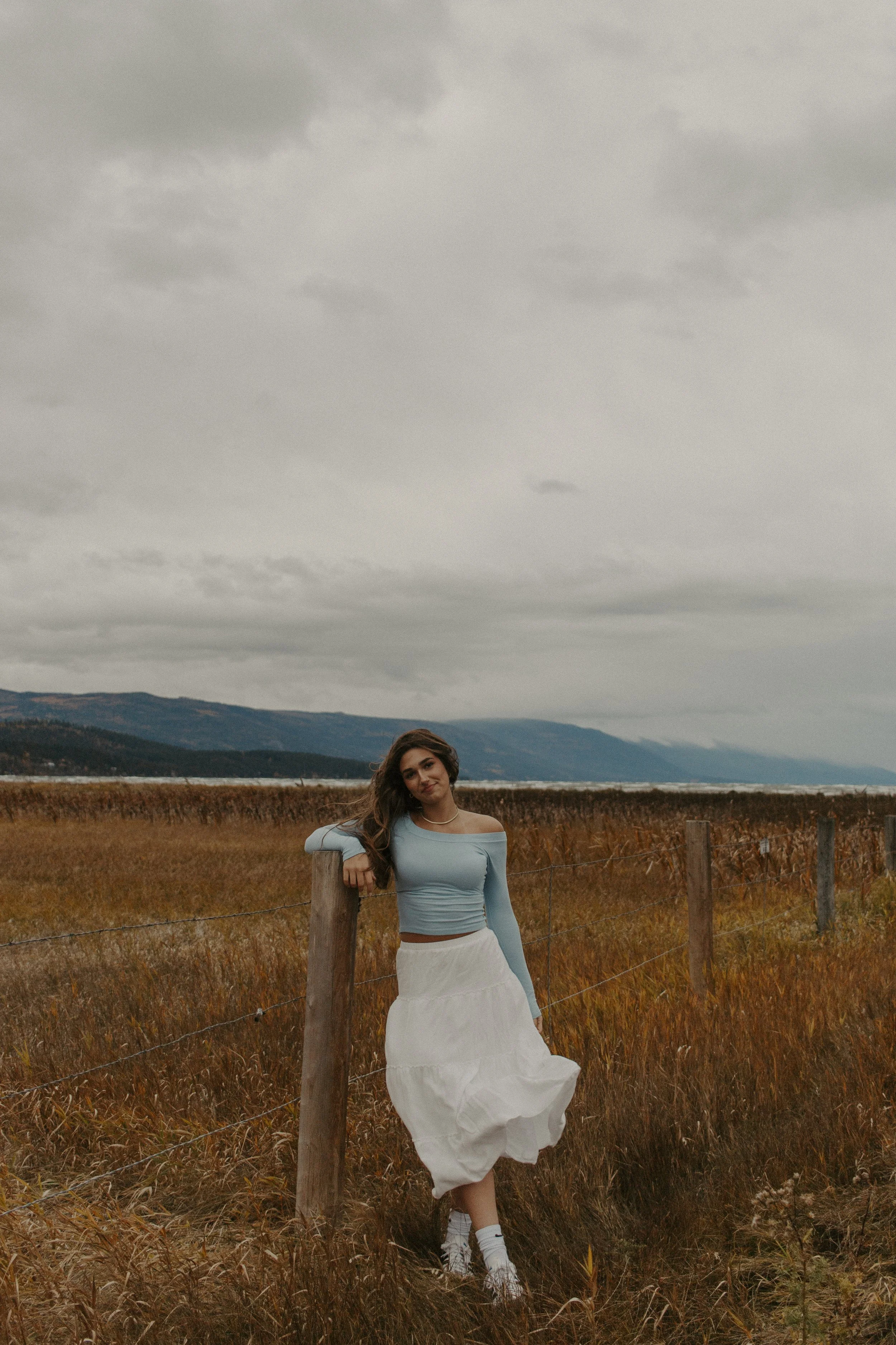 Girl leans against fence in field in front of lake and mountains
