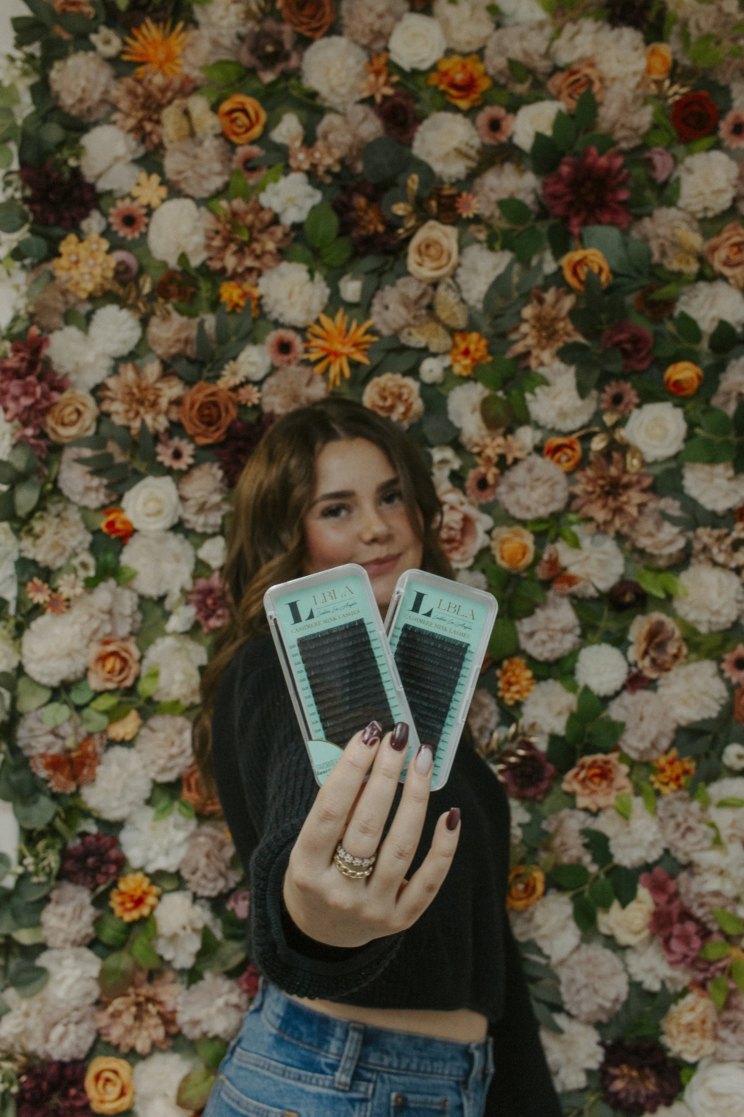 girl holds out arm with eyelash extension packs against flower wall