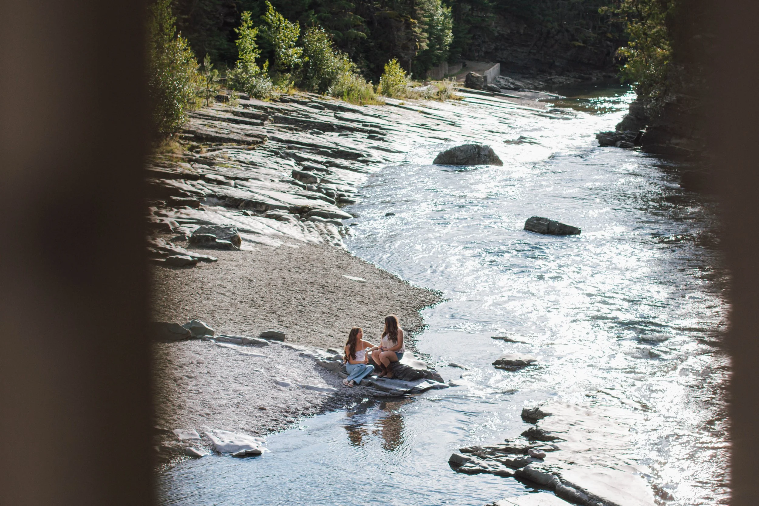 two girls sit on a rock by the river and talk surrounded by trees