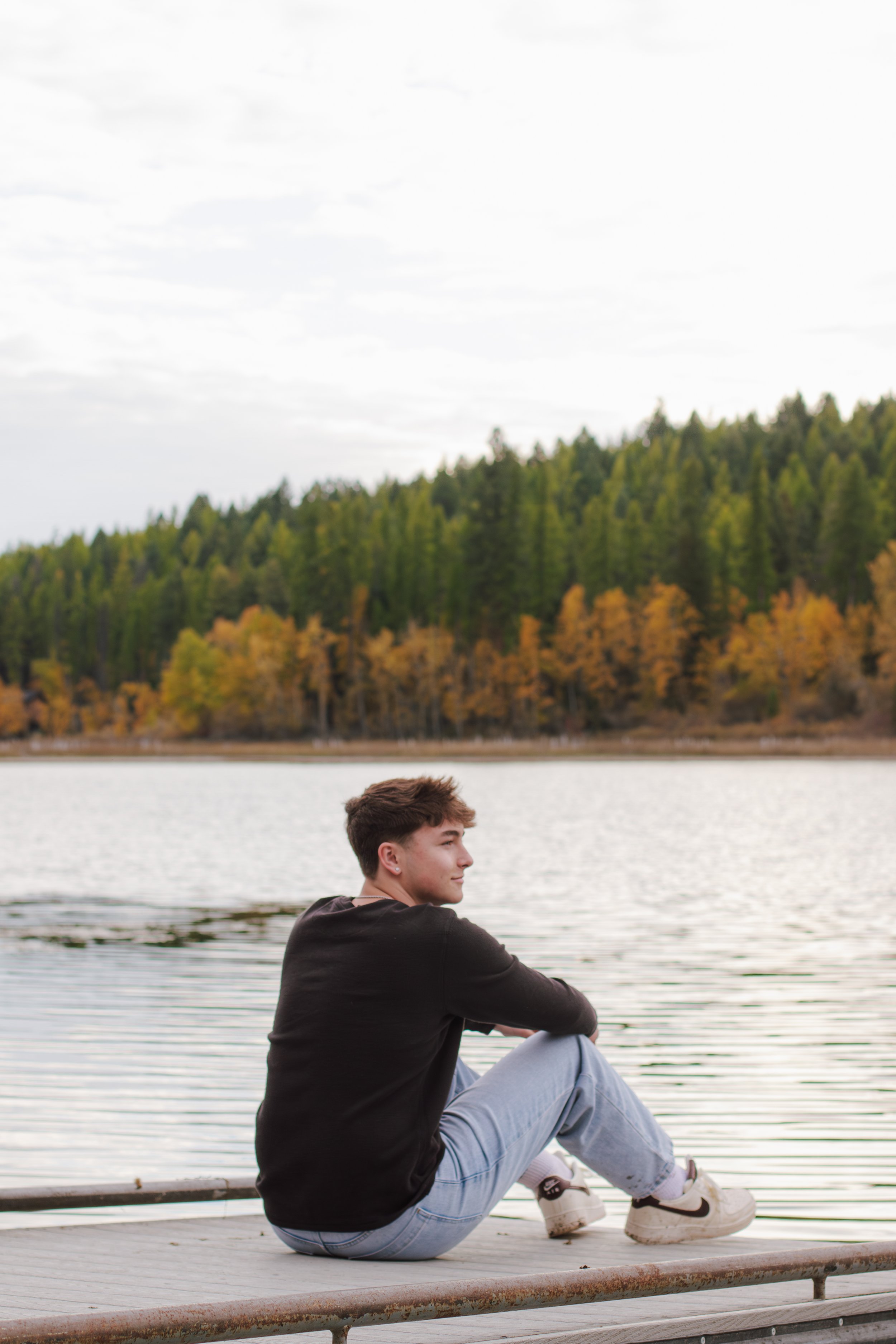 boy sits on end of dock looking out on the lake and fall trees