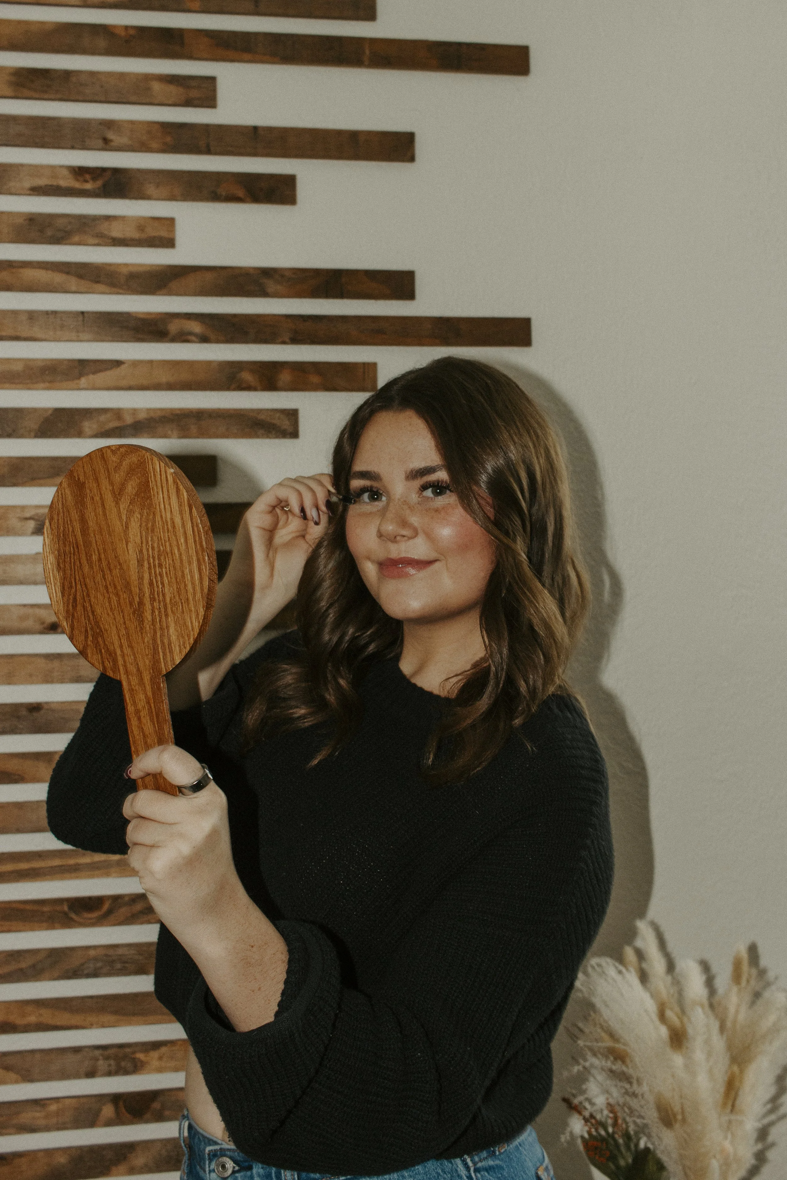girl hold wooden mirror up and brushes her eyelashes