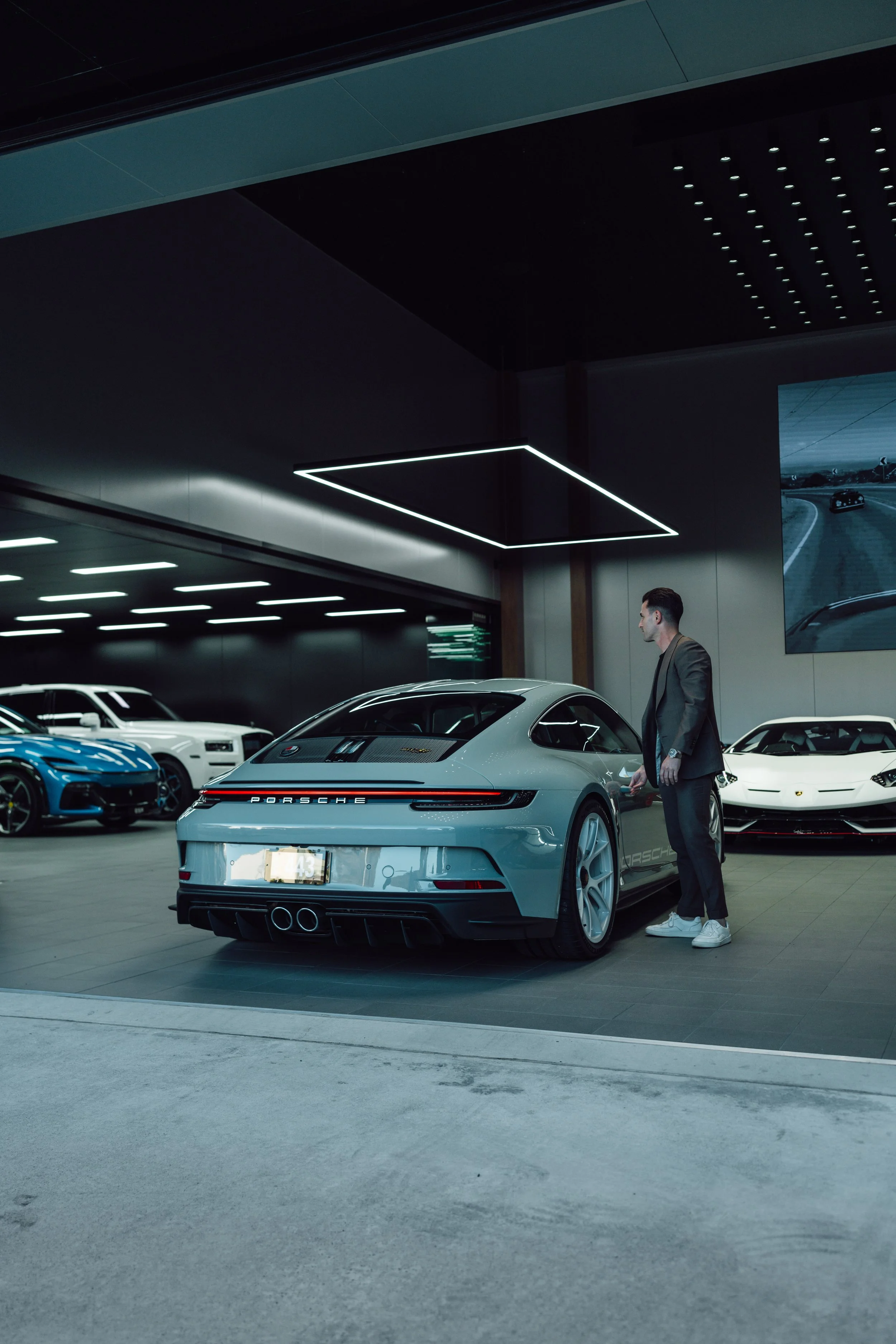 A man in a suit standing next to a gray Porsche sports car in a showroom with luxury and sports cars in the background.
