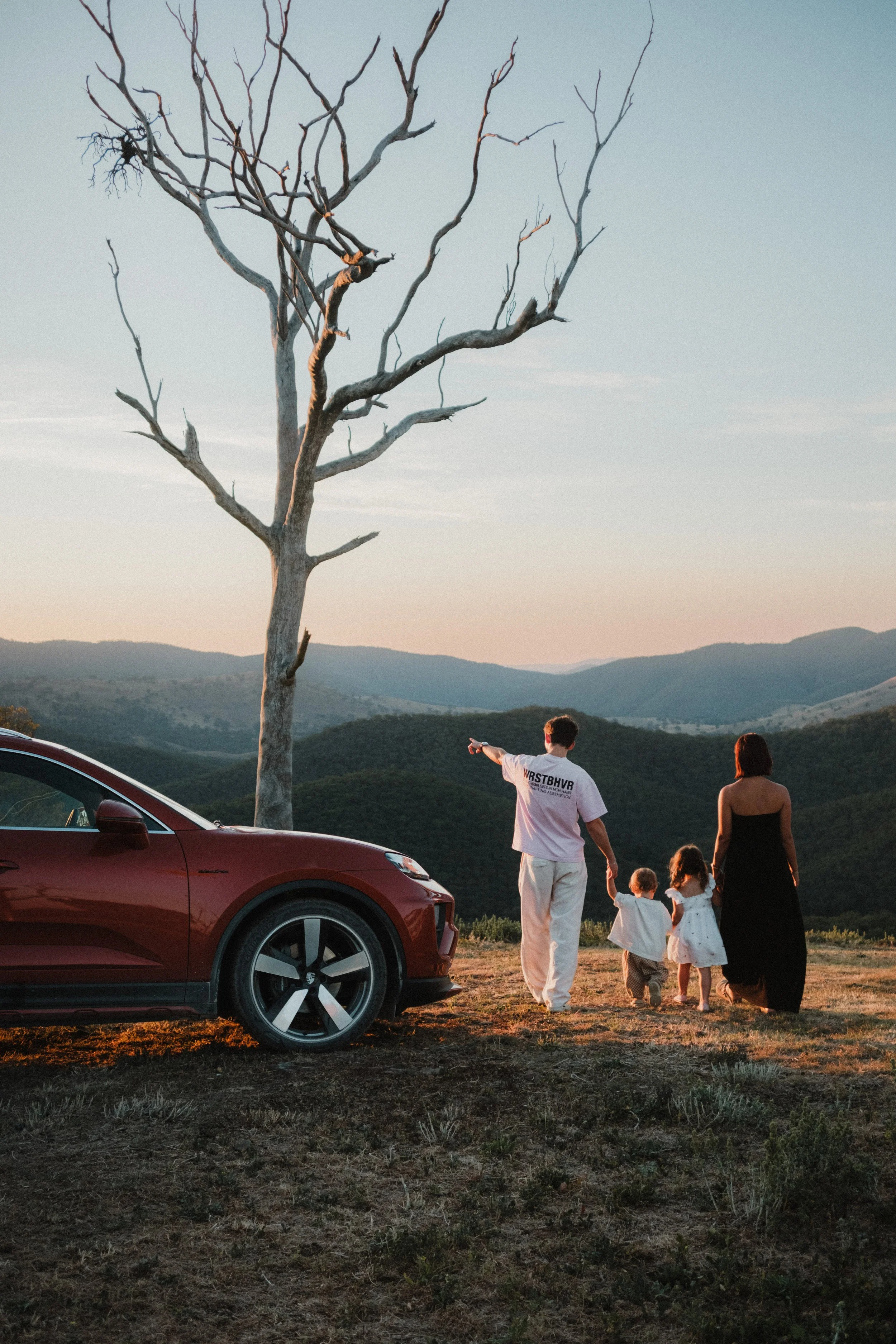 A family of five, including a man, a woman, and three children, walking hand-in-hand away from a red car on grassy terrain, with mountains in the background and a leafless tree nearby during sunset.