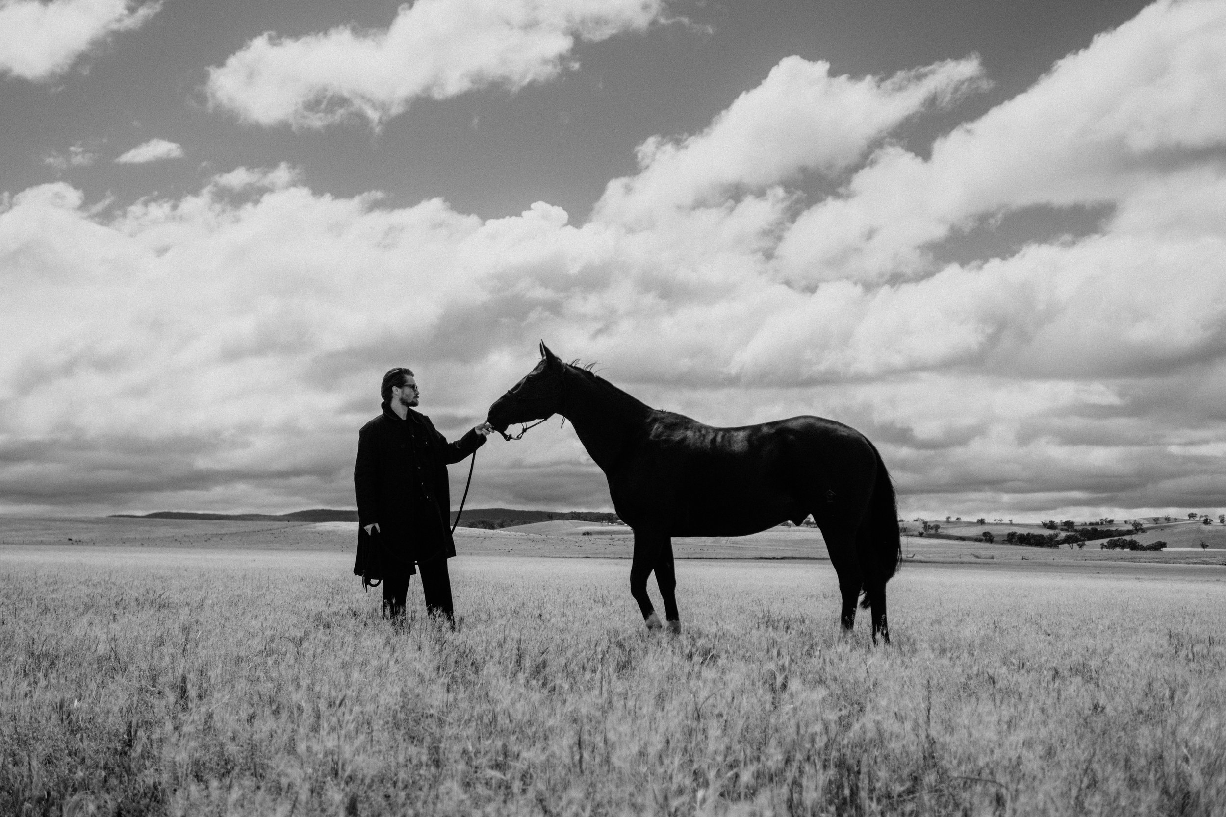 A man in dark clothing holding a black horse's lead in an open field under a cloudy sky.