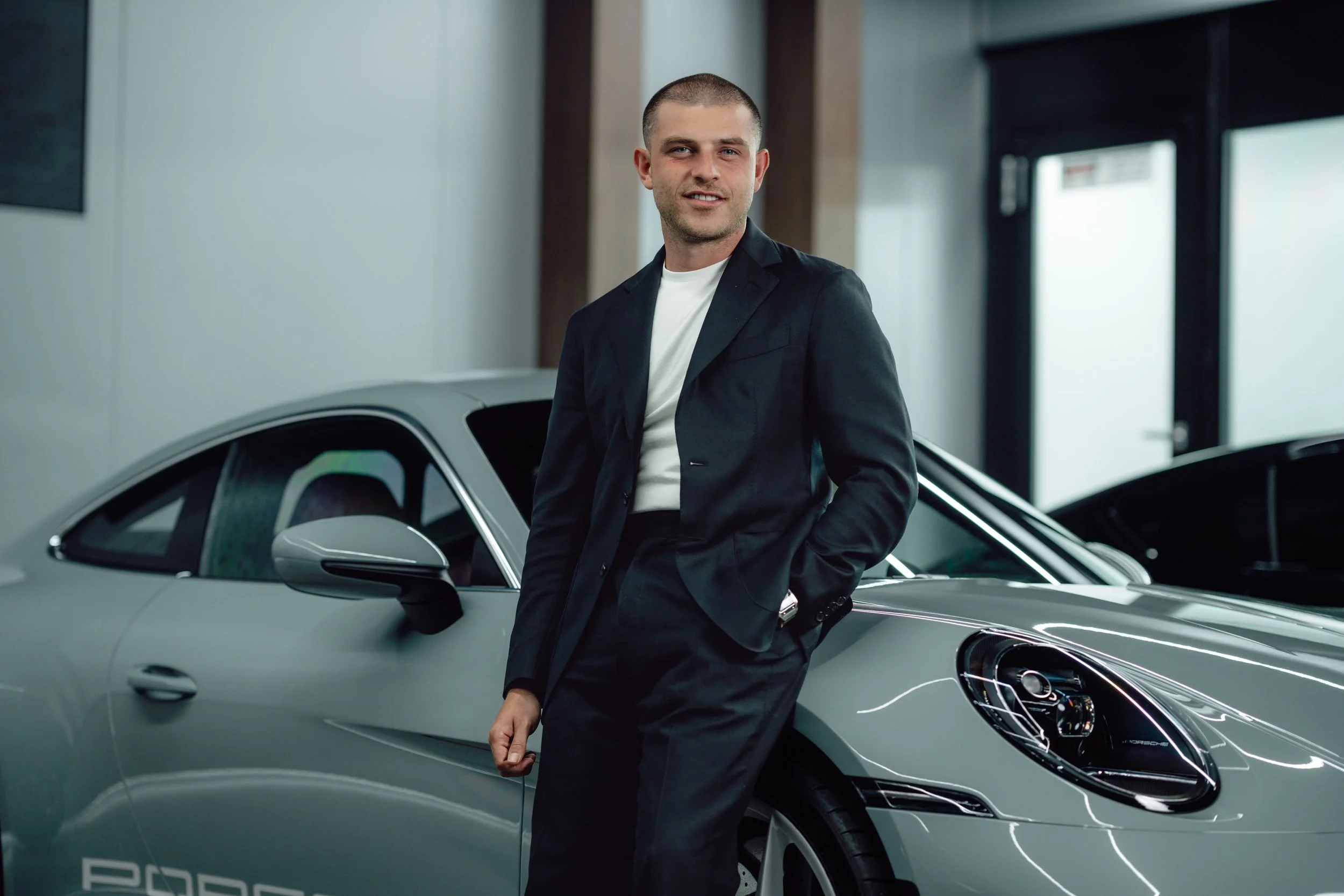 Man in black suit leaning against a silver Porsche car in an indoor showroom.